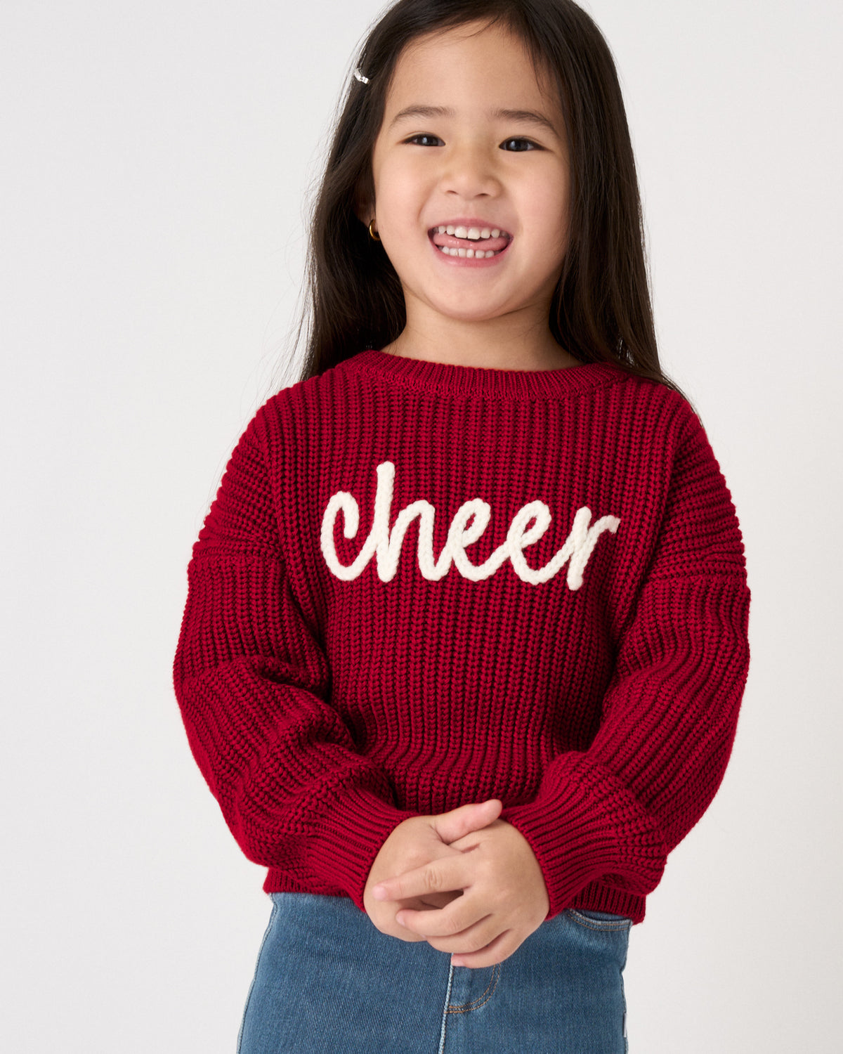 A young girl smiling while wearing a red ribbed knit sweater with the word 'cheer' in white script across the chest, paired with blue jeans, against a plain light gray background.