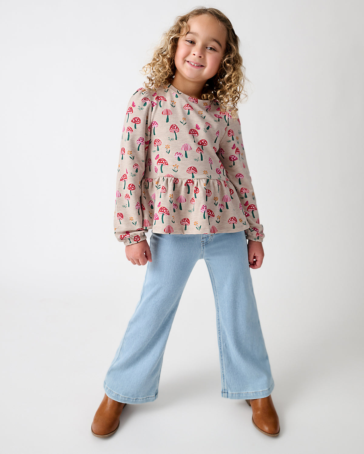 A young girl stands against a light gray studio background, wearing a long-sleeve cream blouse with a pink and green mushroom print, light blue wide-leg jeans, and brown boots.