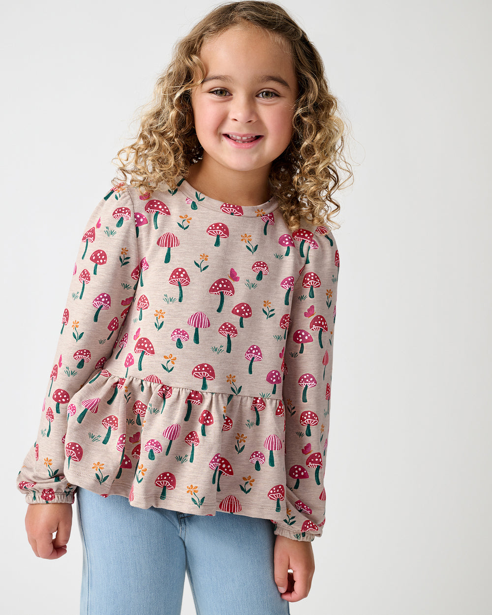 A young girl smiling, wearing a beige long-sleeve top with a colorful mushroom print and light blue jeans, photographed against a solid light gray background.