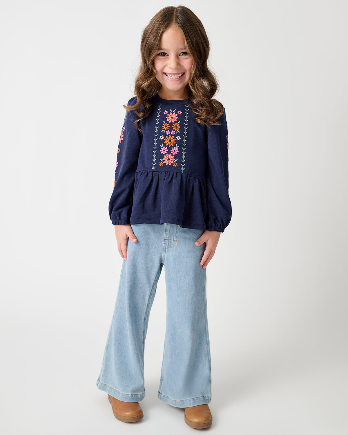 A young girl stands against a light gray studio background, wearing a navy long-sleeve top with colorful floral embroidery on the chest and sleeves, light blue wide-leg jeans, and tan shoes.