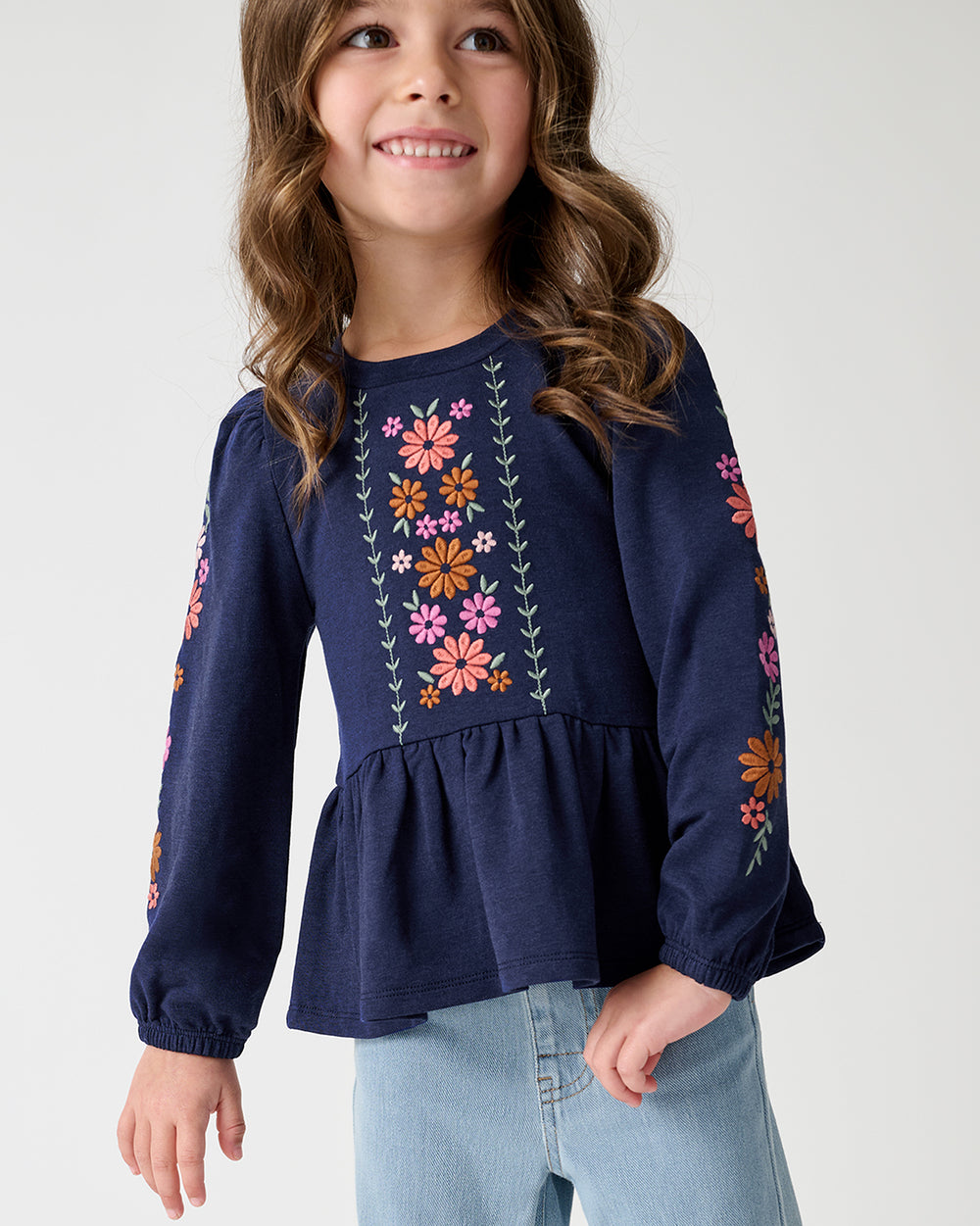 A young girl wearing a navy blue long-sleeve top with colorful floral embroidery and light-wash blue jeans, standing against a light gray studio background.