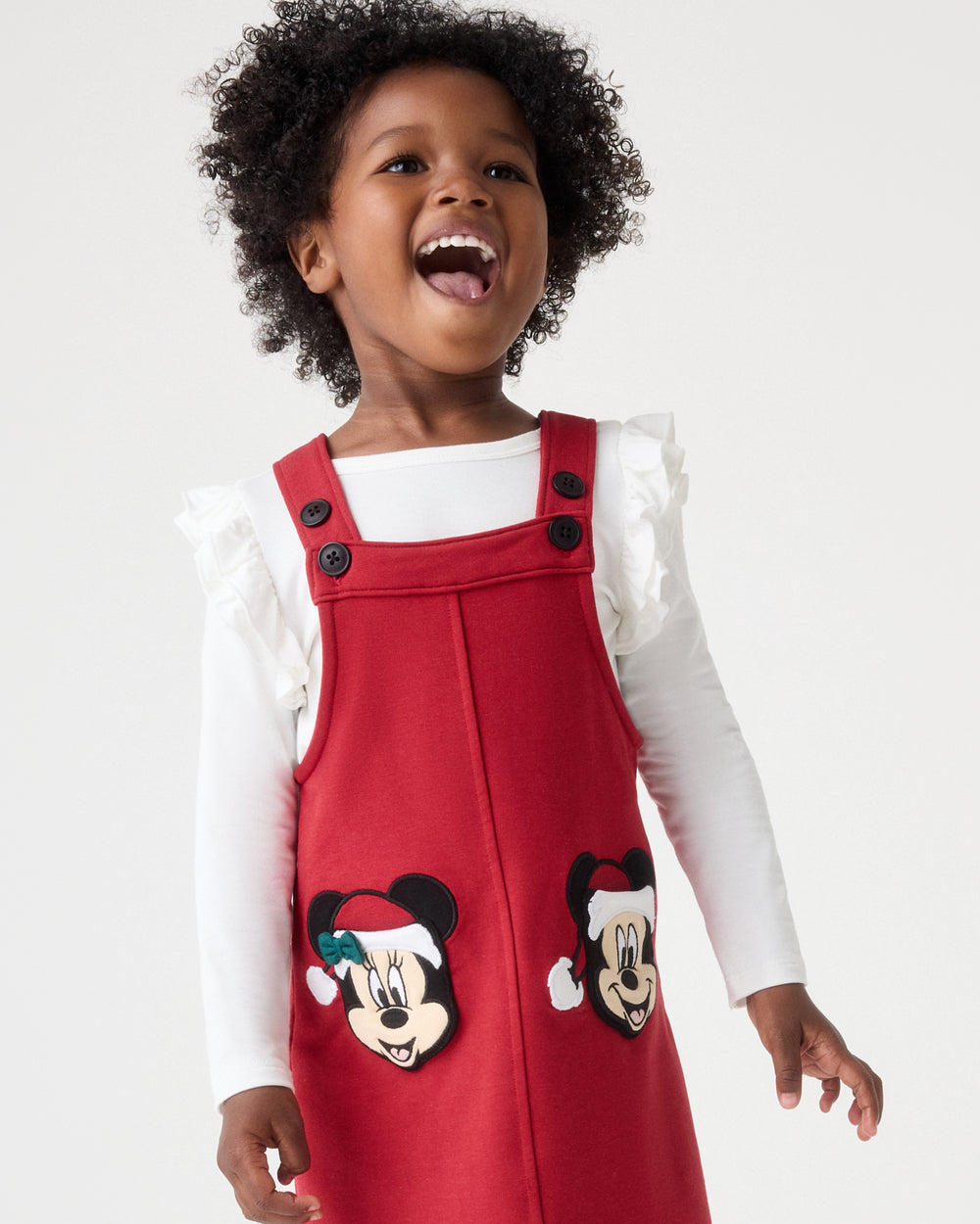 A young child wearing a red holiday-themed pinafore dress over a white long-sleeve ruffled shirt. The dress features embroidered cartoon mouse faces wearing Santa hats on the front pockets, set against a plain light gray studio background.