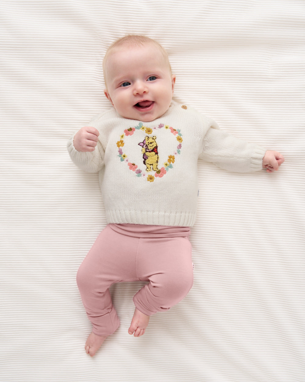 A baby lying on a white textured blanket, wearing a cream knit sweater with a colorful floral heart design and a yellow cartoon character in the center, paired with dusty pink pants.