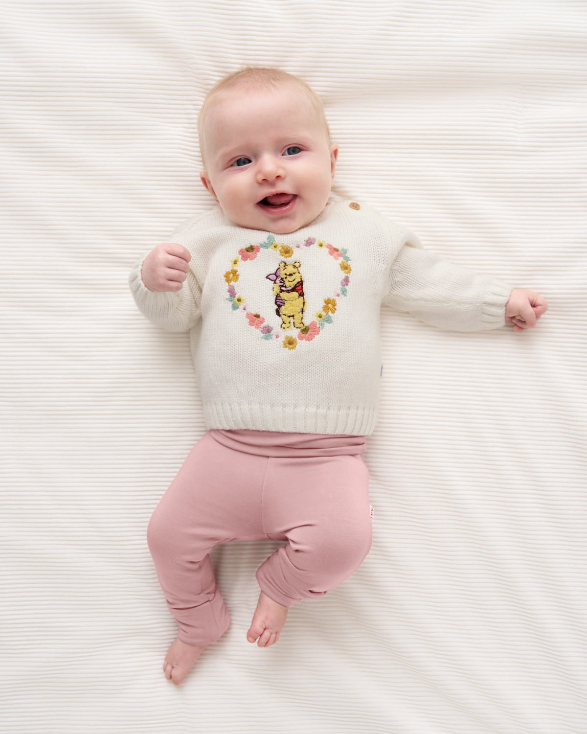 A baby lying on a white textured blanket, wearing a cream knit sweater with a colorful floral heart design and a yellow cartoon character in the center, paired with dusty pink pants.