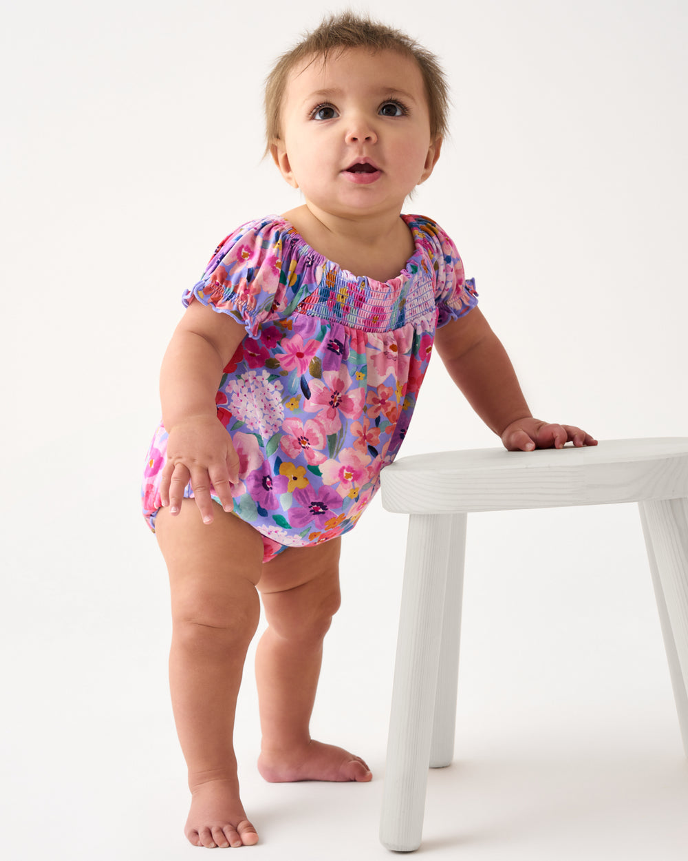Baby wearing the Sweet Sprigs Smocked Bubble Romper holding onto a white stool