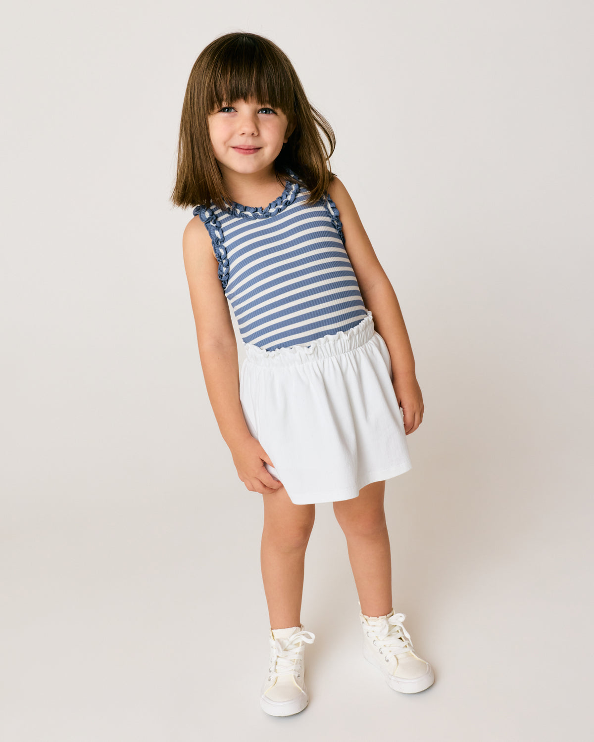 Standing girl wearing the Infinite Blue Stripe Ribbed Ruffle Tank with a white denim skort