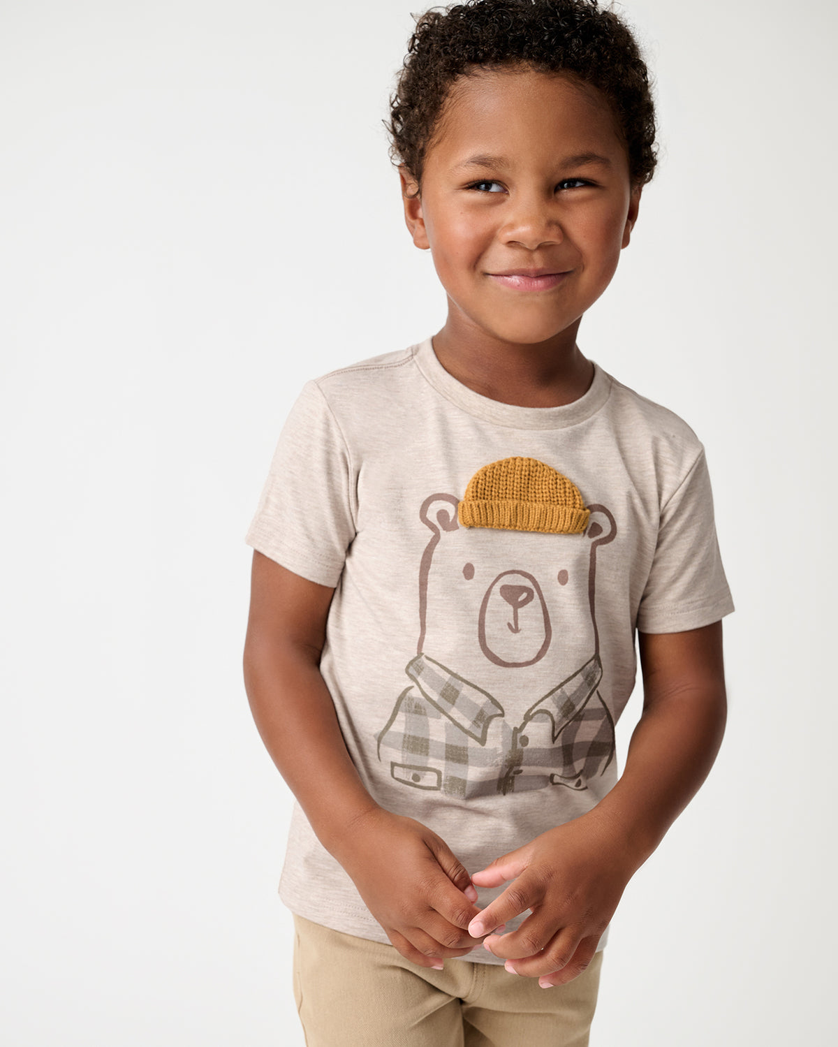 A young child wearing a light beige short-sleeve t-shirt with a cartoon bear graphic wearing a mustard yellow knit hat and plaid shirt. The child is also wearing khaki pants, standing against a clean white studio background.