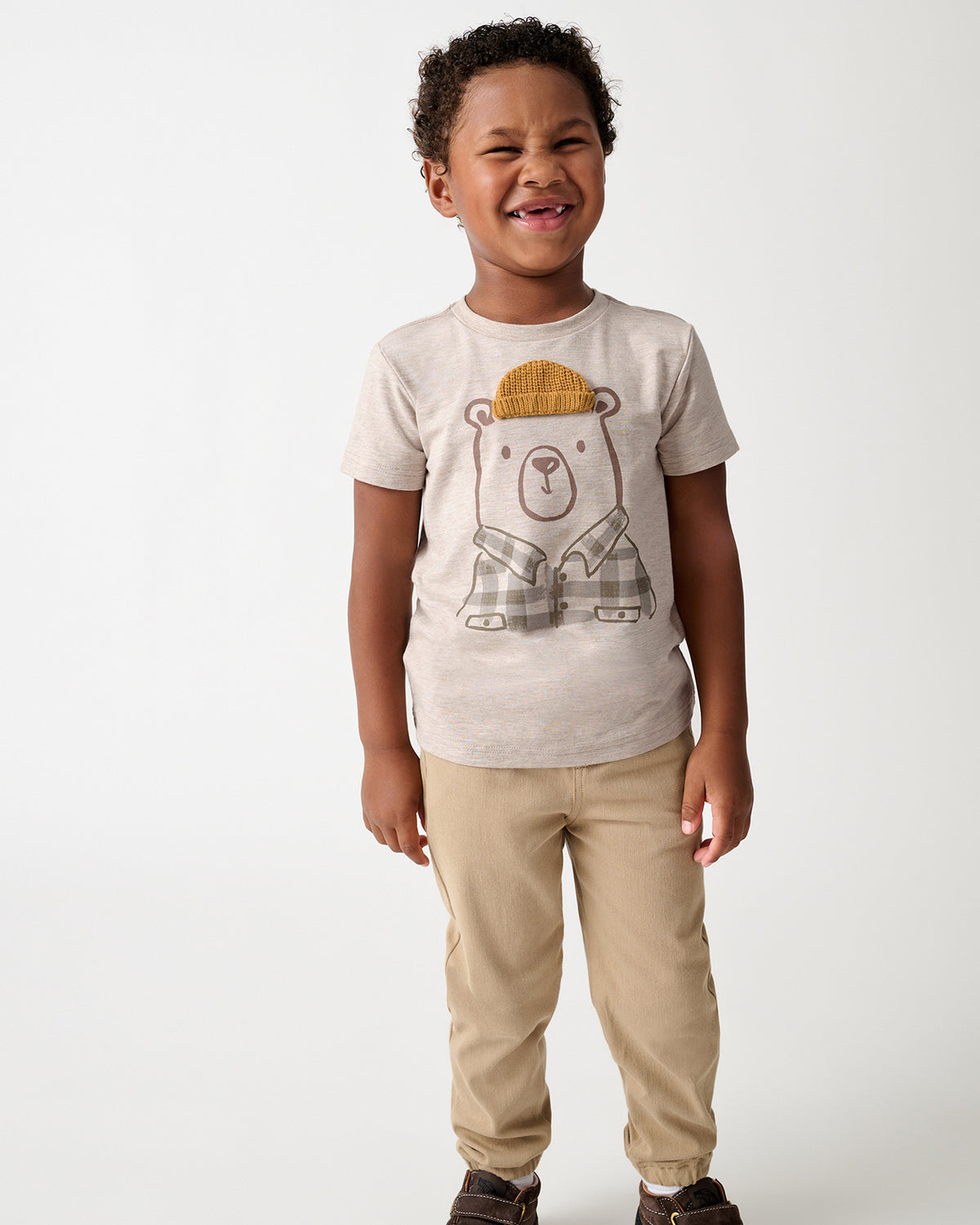 A young child wearing a light beige graphic t-shirt with a cartoon bear wearing a yellow knit hat and plaid shirt, paired with khaki pants and brown shoes, standing against a plain light gray background.