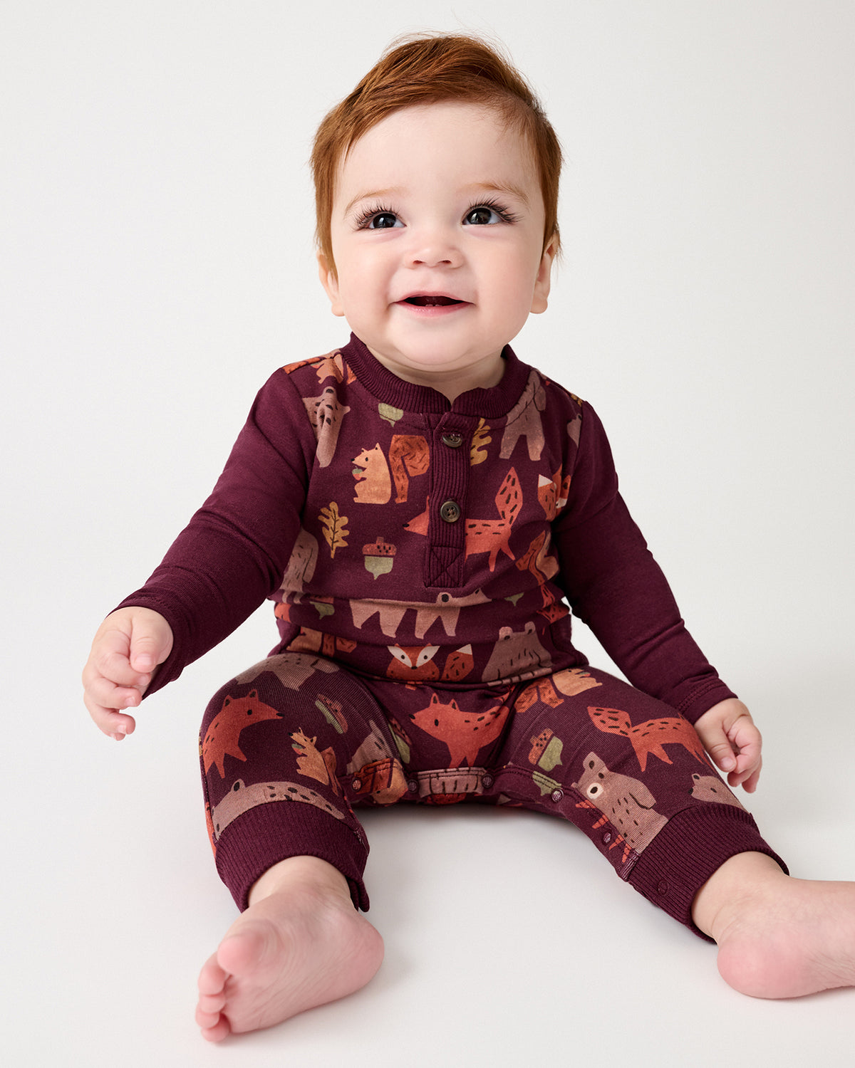 A smiling baby sitting on a white studio floor wearing a burgundy long-sleeve pajama top and matching pants with an autumn forest print of foxes, leaves, and acorns.