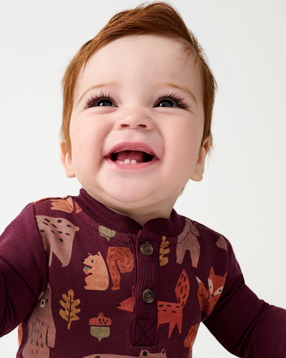 A close-up of a smiling baby wearing a burgundy long-sleeve henley romper with a woodland animal print including foxes, squirrels, and acorns, set against a plain light gray background.