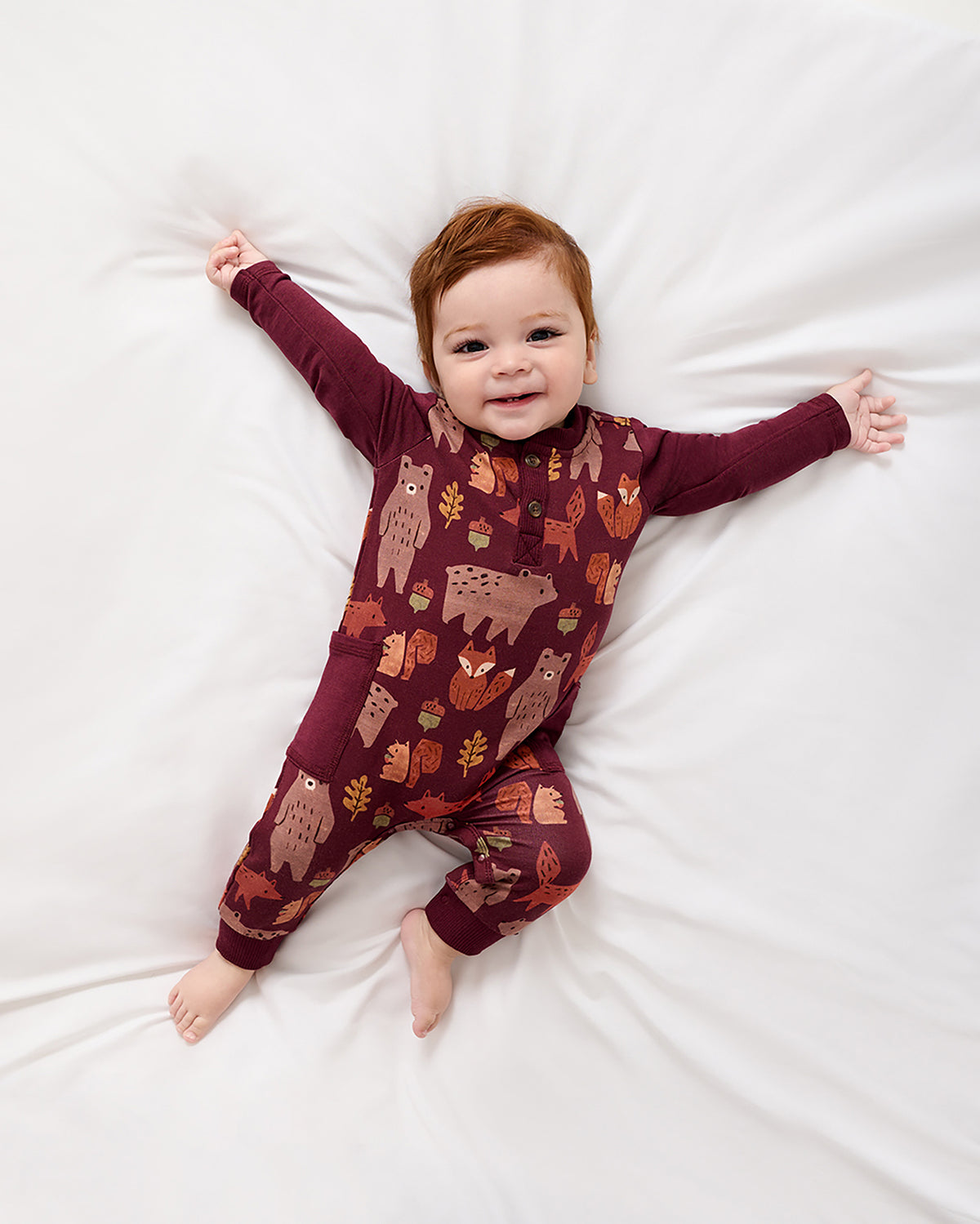 A smiling baby lies on a white bed wearing a burgundy long-sleeve footless one-piece pajama with an autumn animal print of owls, foxes, and leaves.