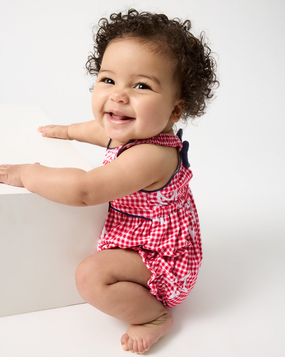 Crouching child wearing the Celebration Gingham Bow Back Bubble Romper