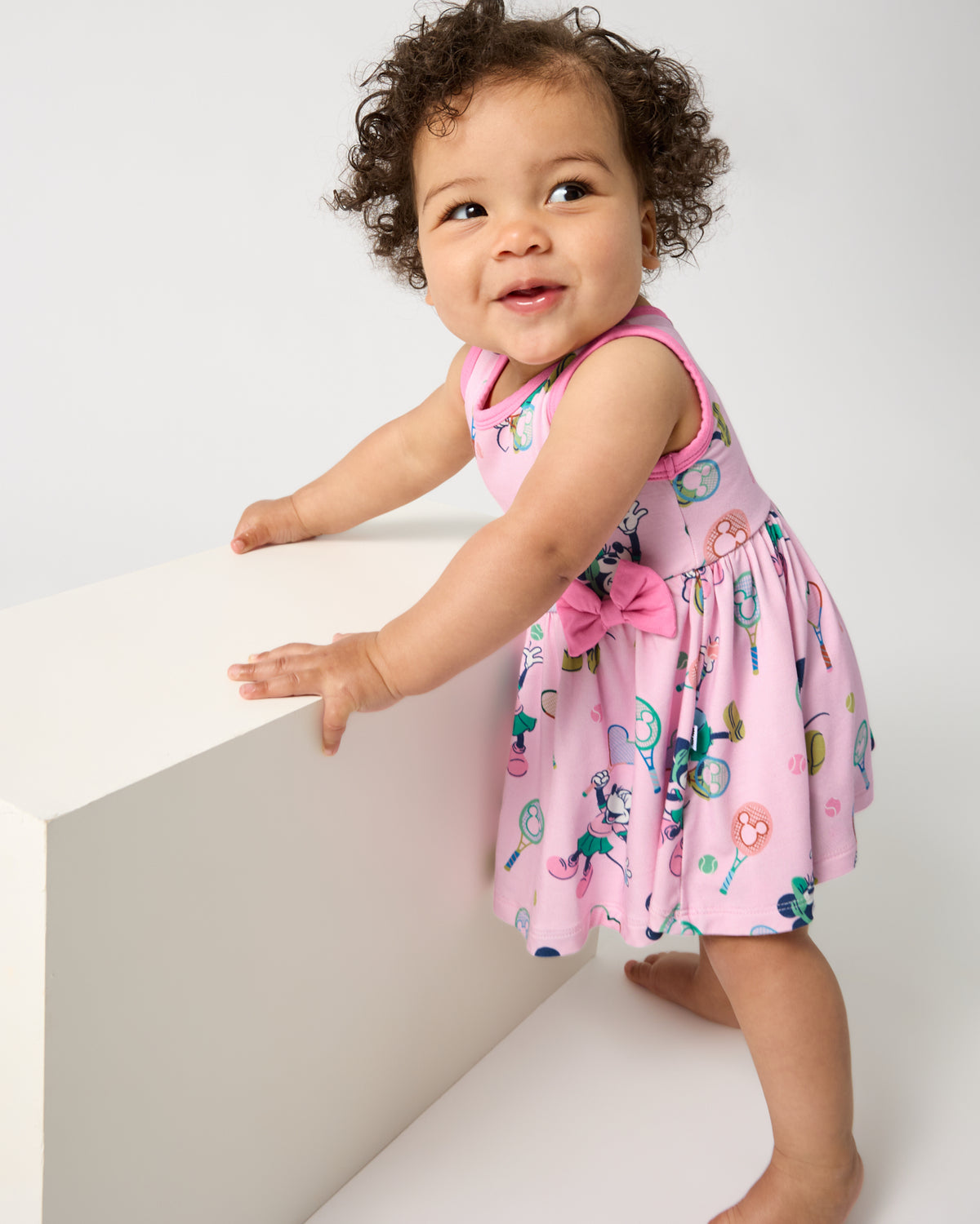 Baby wearing the Disney Mickey Mouse & Minnie Mouse Match Twirl Dress with Bodysuit holding onto a wooden table