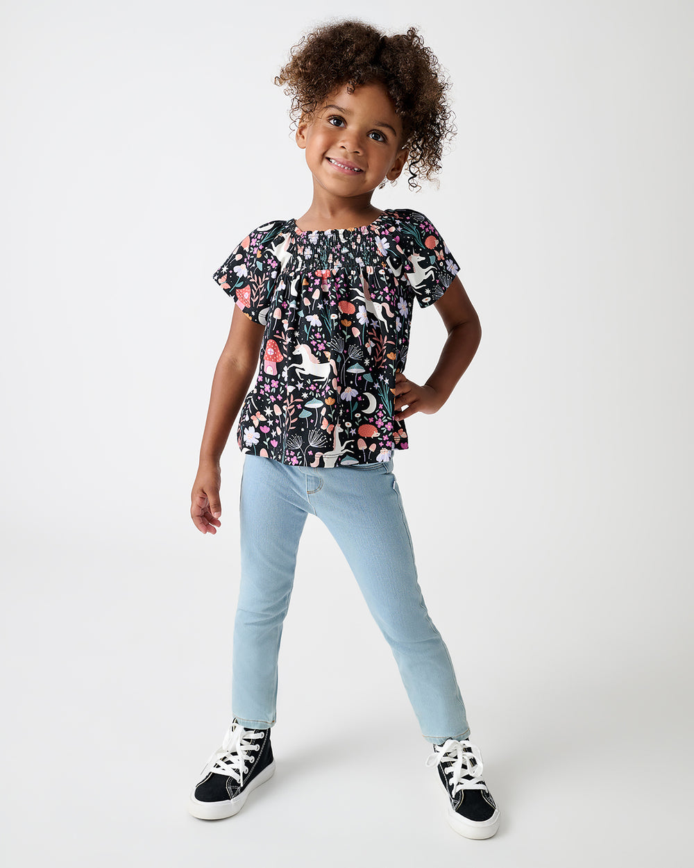 A young girl stands against a light gray studio background wearing a short-sleeve ruffled blouse with a multicolor floral and butterfly pattern, light blue jeans, and black sneakers.