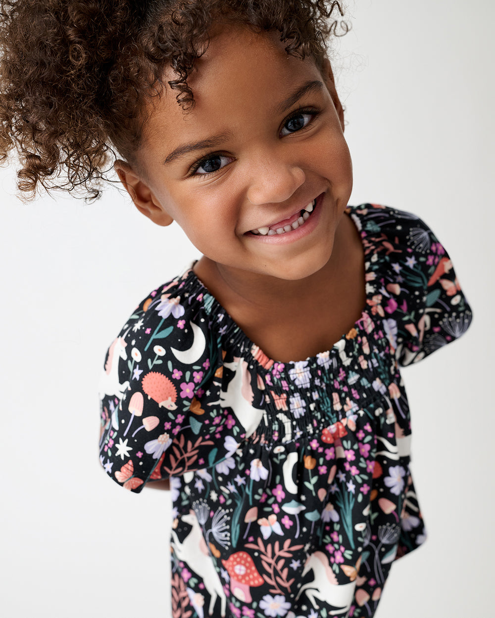 A smiling young girl with curly brown hair wearing a short-sleeve smocked top with a colorful woodland print of flowers, mushrooms, and white birds on a black background, photographed against a clean white studio backdrop.