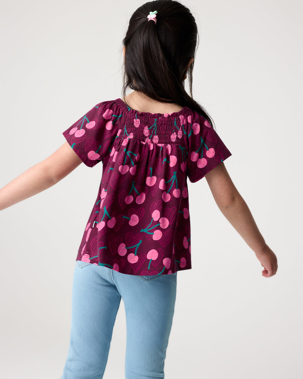 Back view of girl wearing the Cherry Charm Smocked Flutter Top and Denim Pants