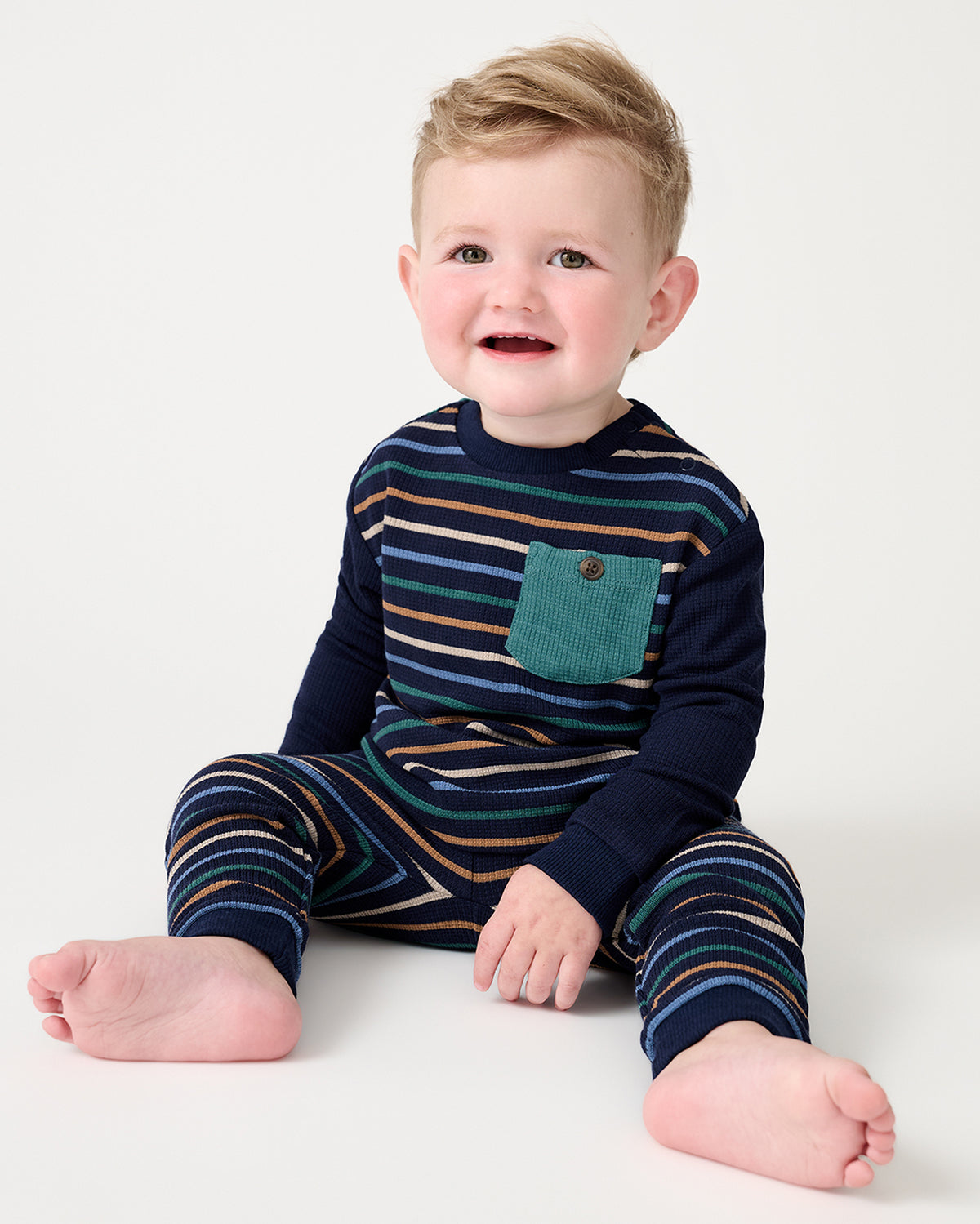 A smiling toddler sitting on a white studio floor wearing a navy long-sleeve jogger set with thin multicolor stripes and a teal chest pocket.