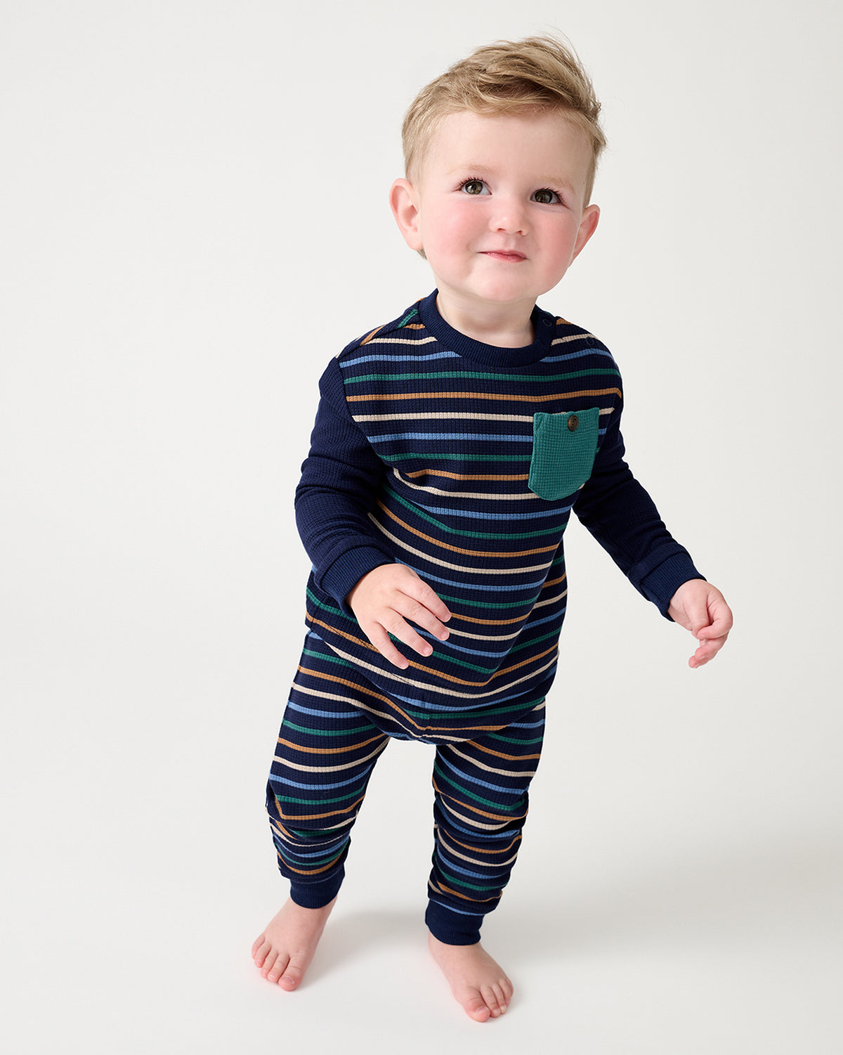 A toddler standing barefoot against a light gray studio background, wearing a long-sleeve striped two-piece set with a teal chest pocket.