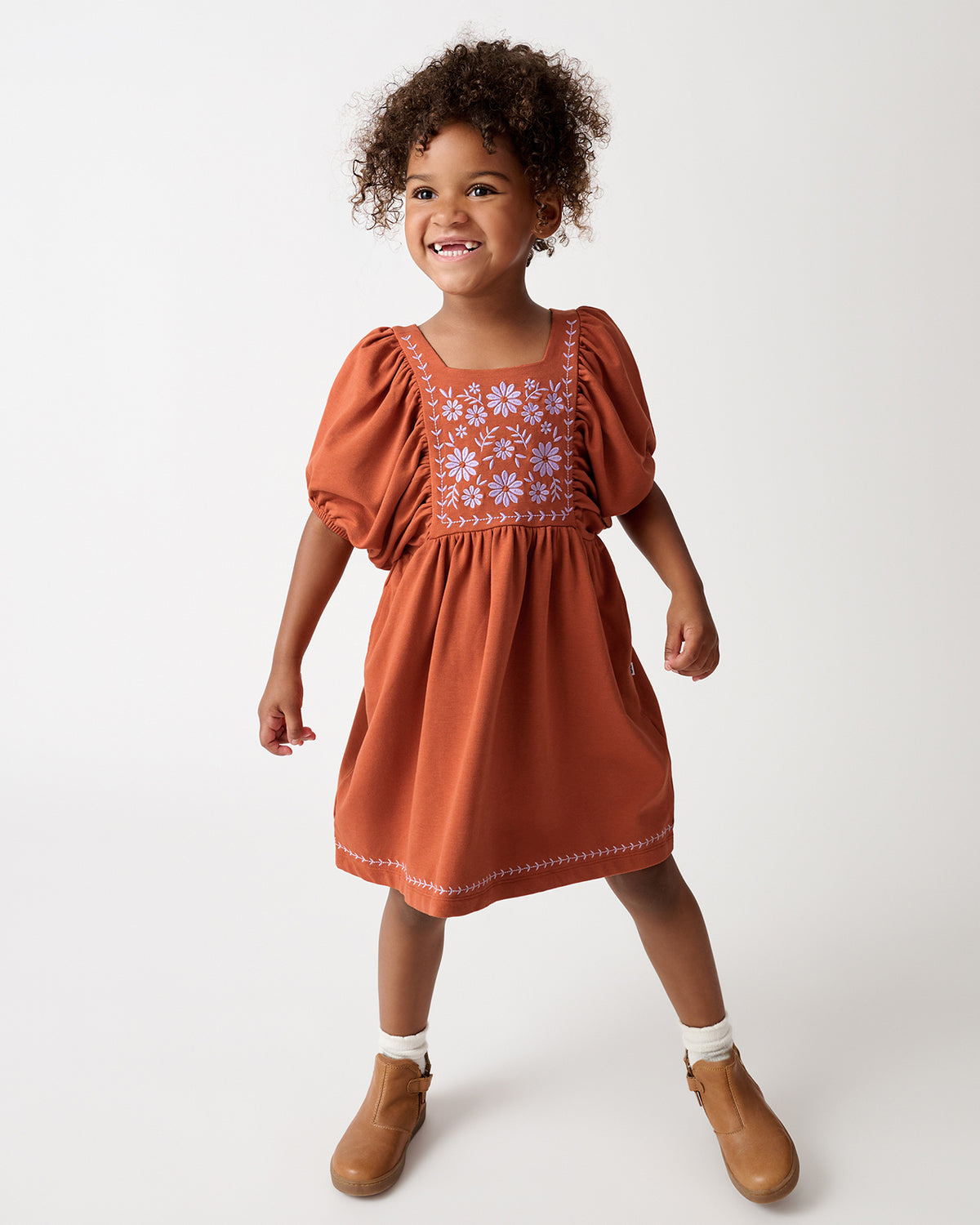A young child wearing a terracotta orange dress with floral embroidery on the bodice, styled with white socks and tan boots, standing against a light gray studio background.
