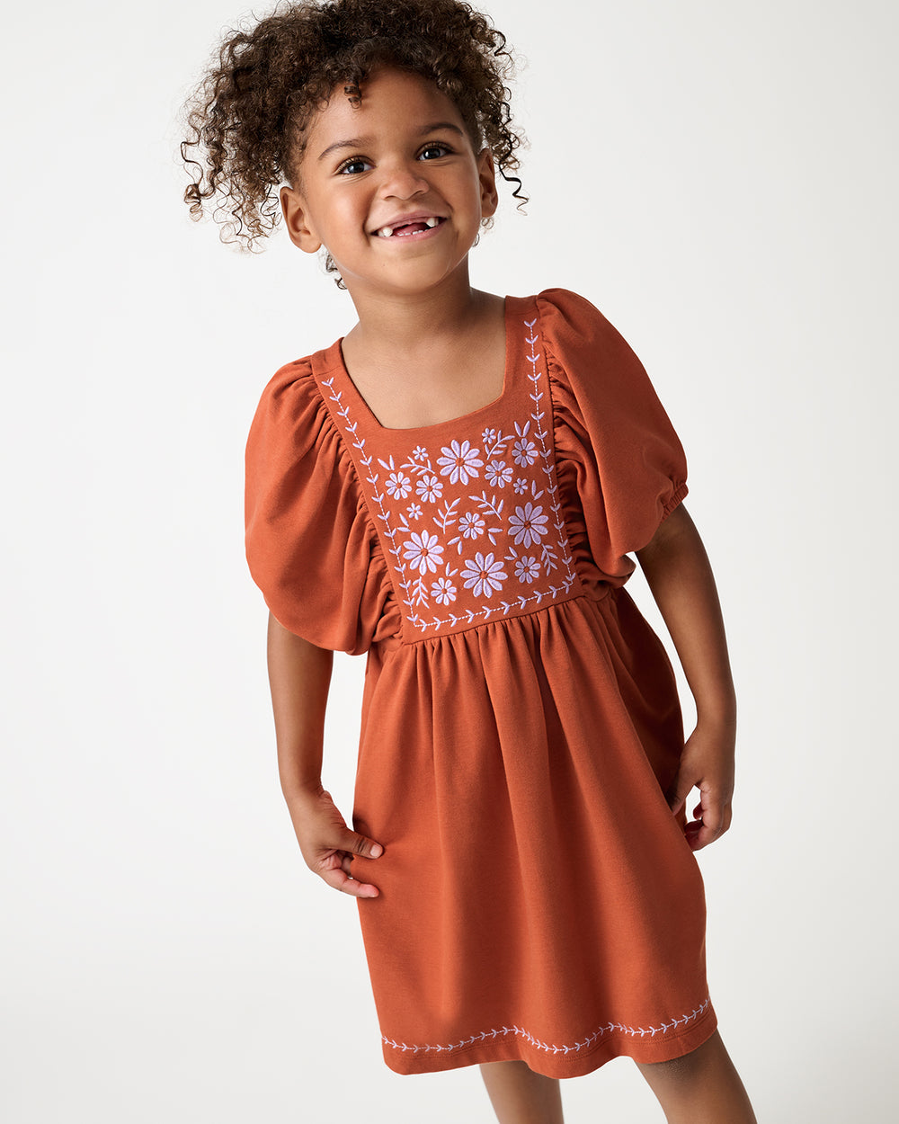 A young child with curly brown hair wearing a rust-orange dress with floral embroidery on the bodice, photographed against a plain light gray studio background.
