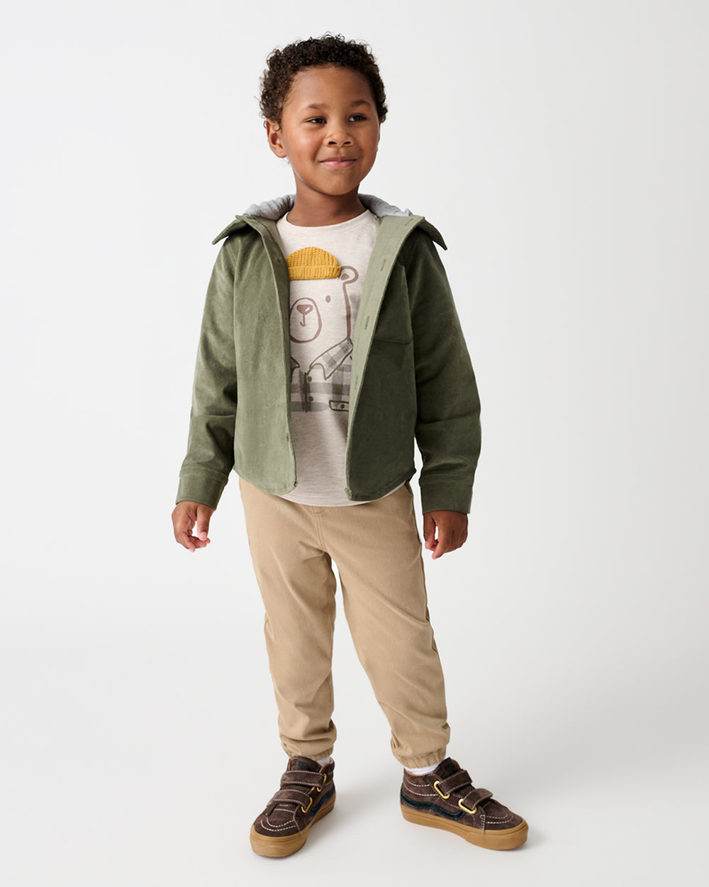 A young boy modeling a green hooded jacket over a graphic t-shirt, paired with beige pants and brown sneakers, against a plain light gray studio background.