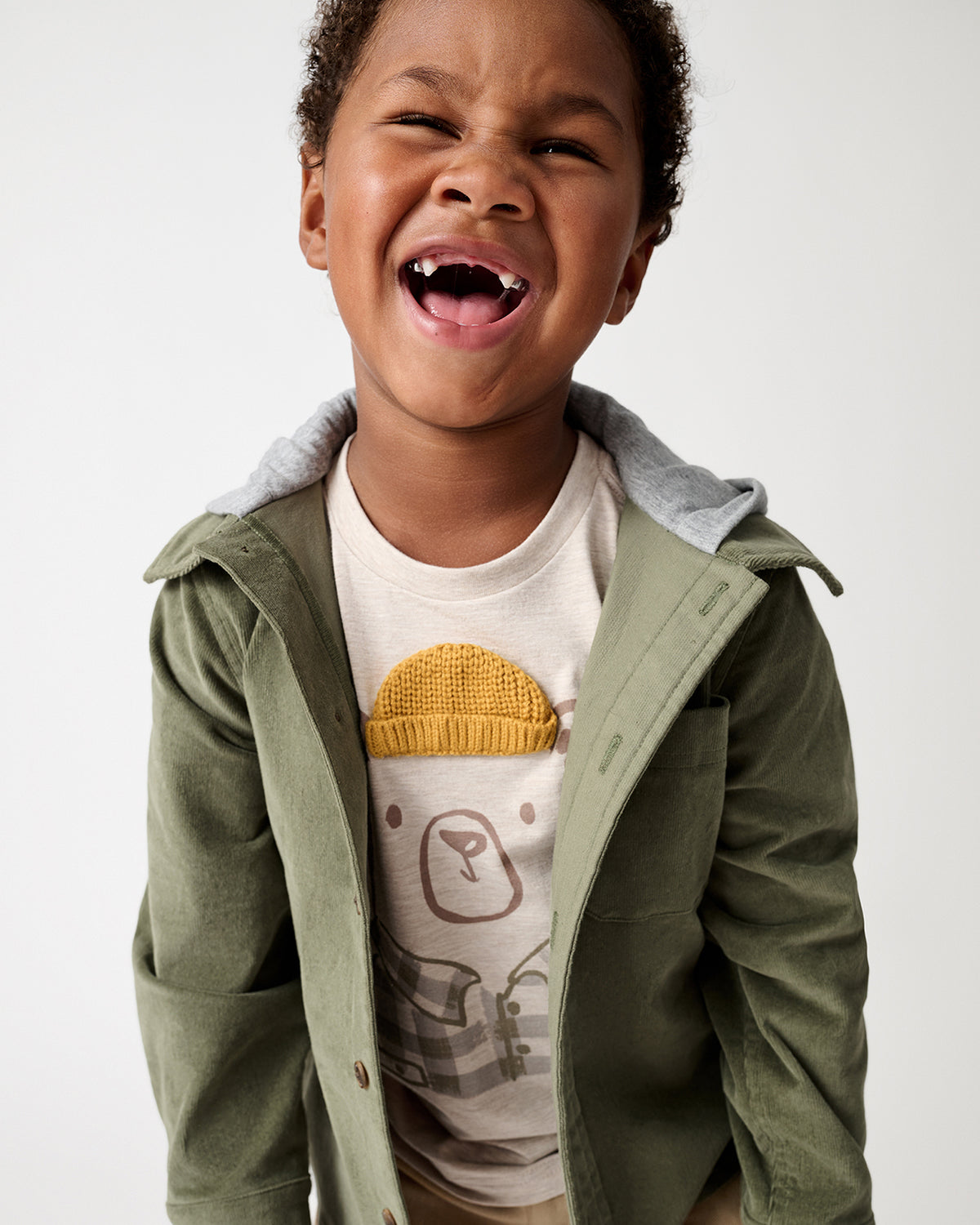 A young child wearing an olive green hooded jacket over a cream graphic t-shirt, captured in a close-up shot against a light gray background.
