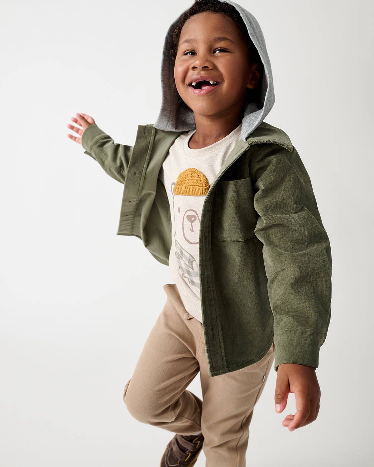 A young child wearing an olive green corduroy hooded jacket over a white graphic t-shirt and beige pants, captured mid-jump against a clean white studio background.