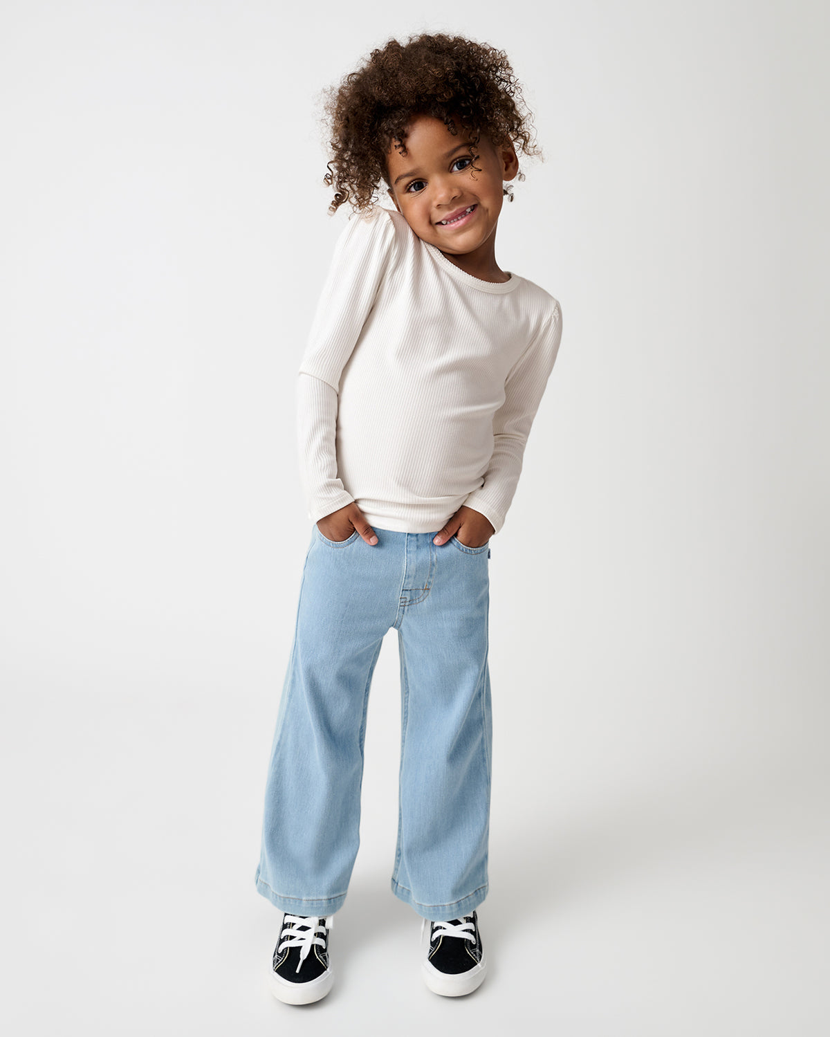 A girl stands against a light gray studio background, wearing a cream long-sleeve top, light blue wide-leg jeans, and black sneakers.