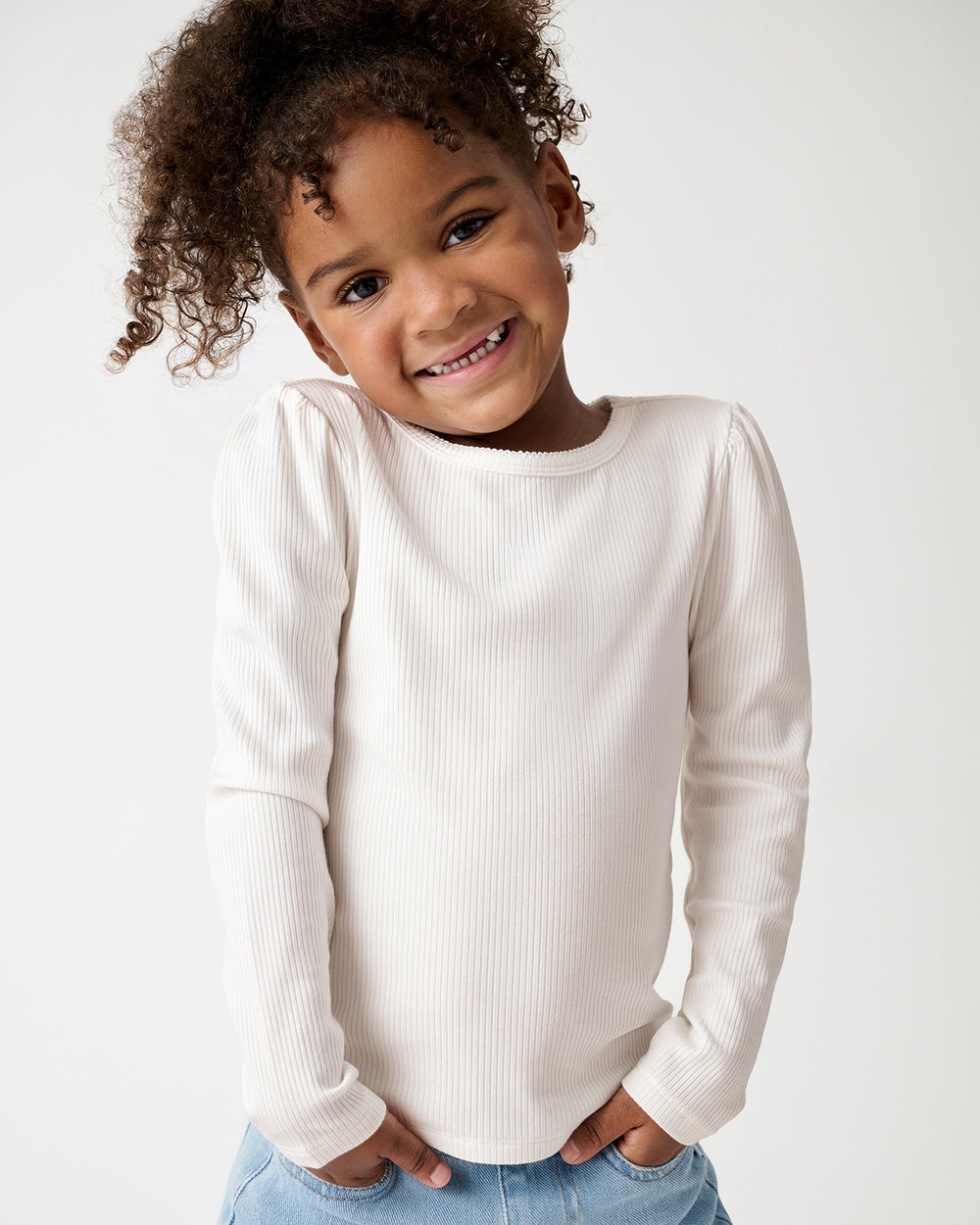 A smiling young girl wearing a cream-colored ribbed long-sleeve top and light blue jeans, posing against a plain white studio background.