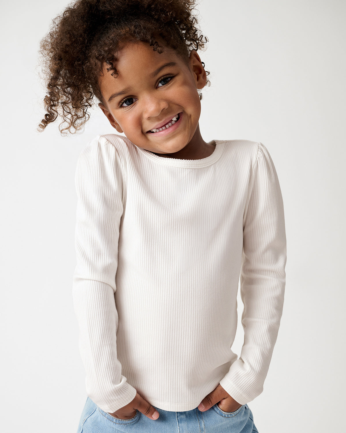 A smiling young girl wearing a cream-colored ribbed long-sleeve top and light blue jeans, posing against a plain white studio background.