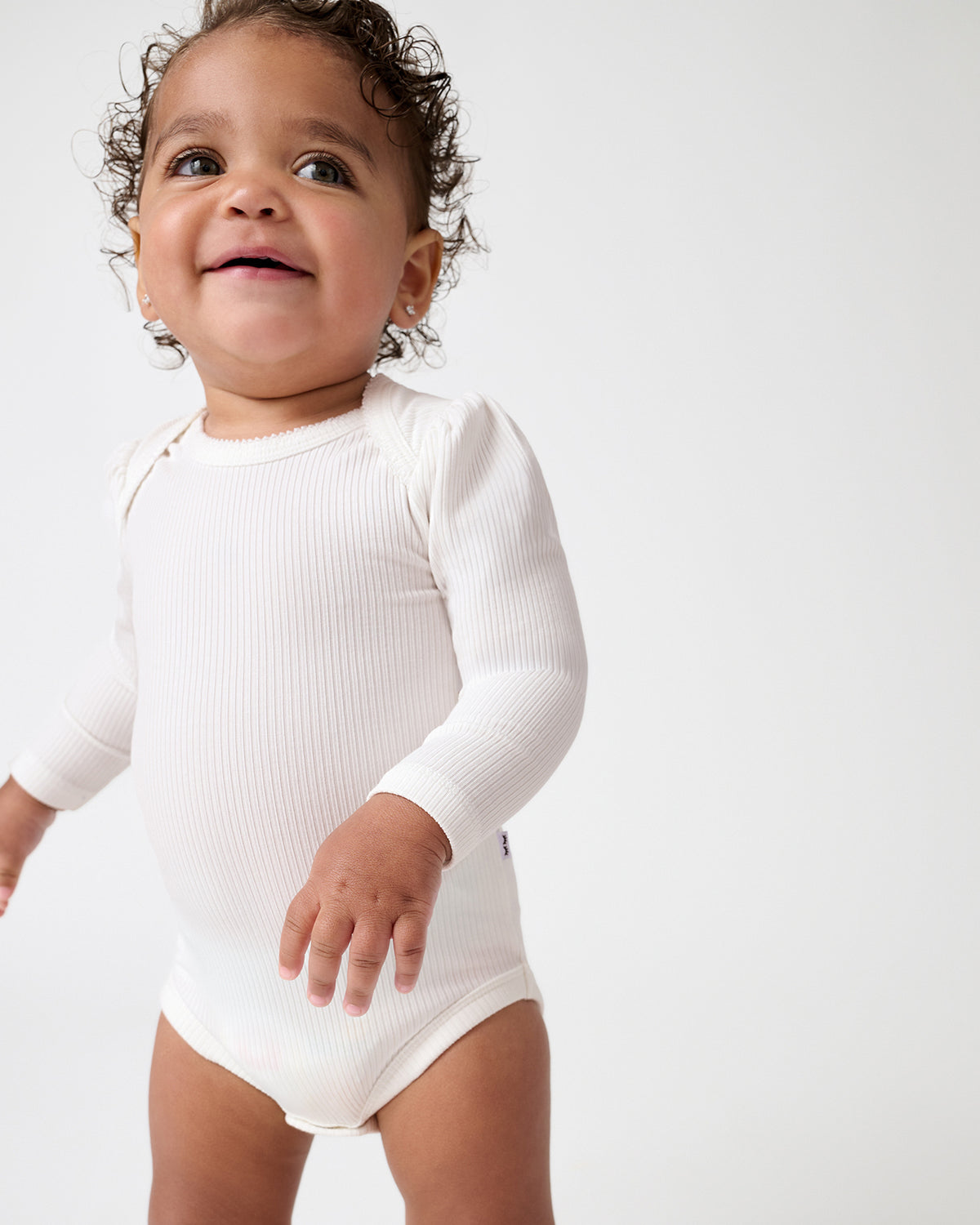 A smiling baby wearing a white ribbed long-sleeve bodysuit, standing against a plain light gray studio background.