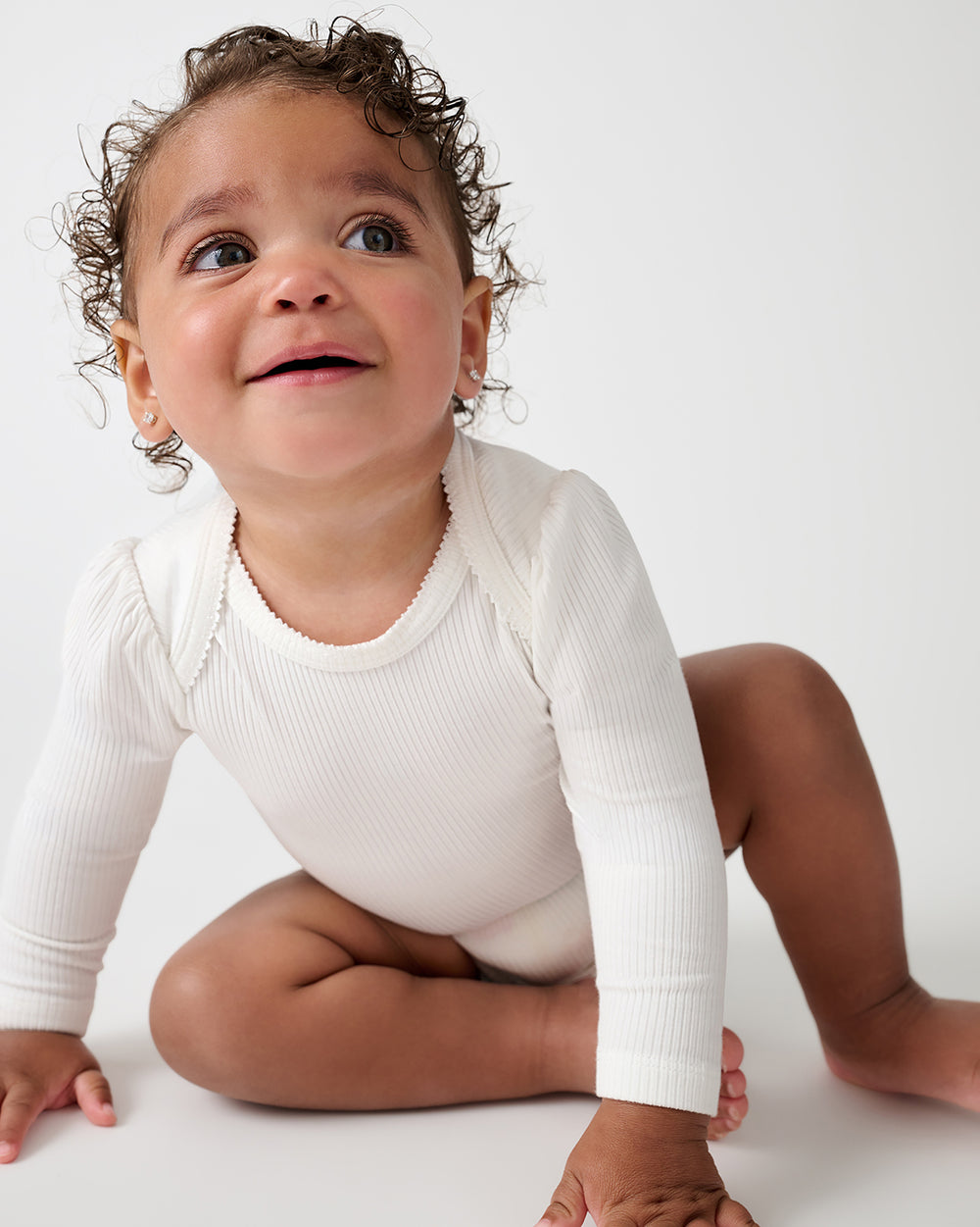 A baby wearing a white long-sleeve ribbed bodysuit, sitting on a white studio floor and looking upward with a smile.