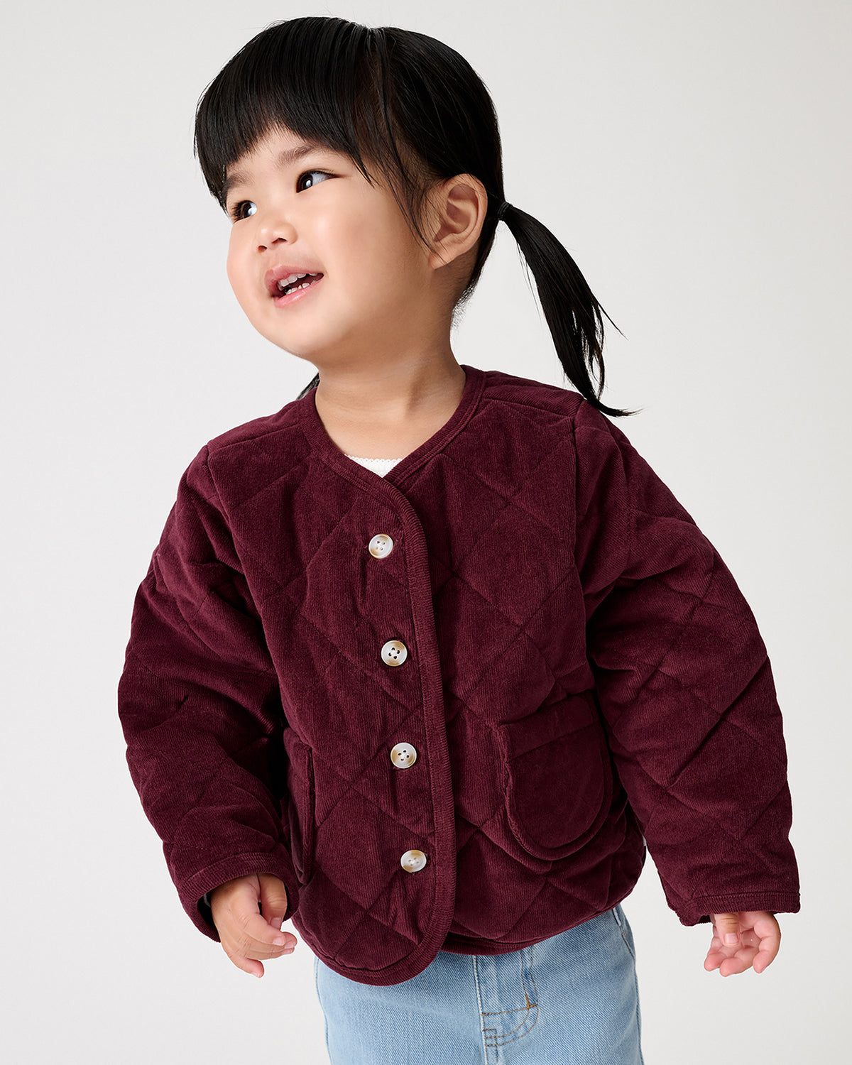 A young child wearing a burgundy quilted jacket with buttons, paired with light blue jeans, photographed against a plain light gray background.