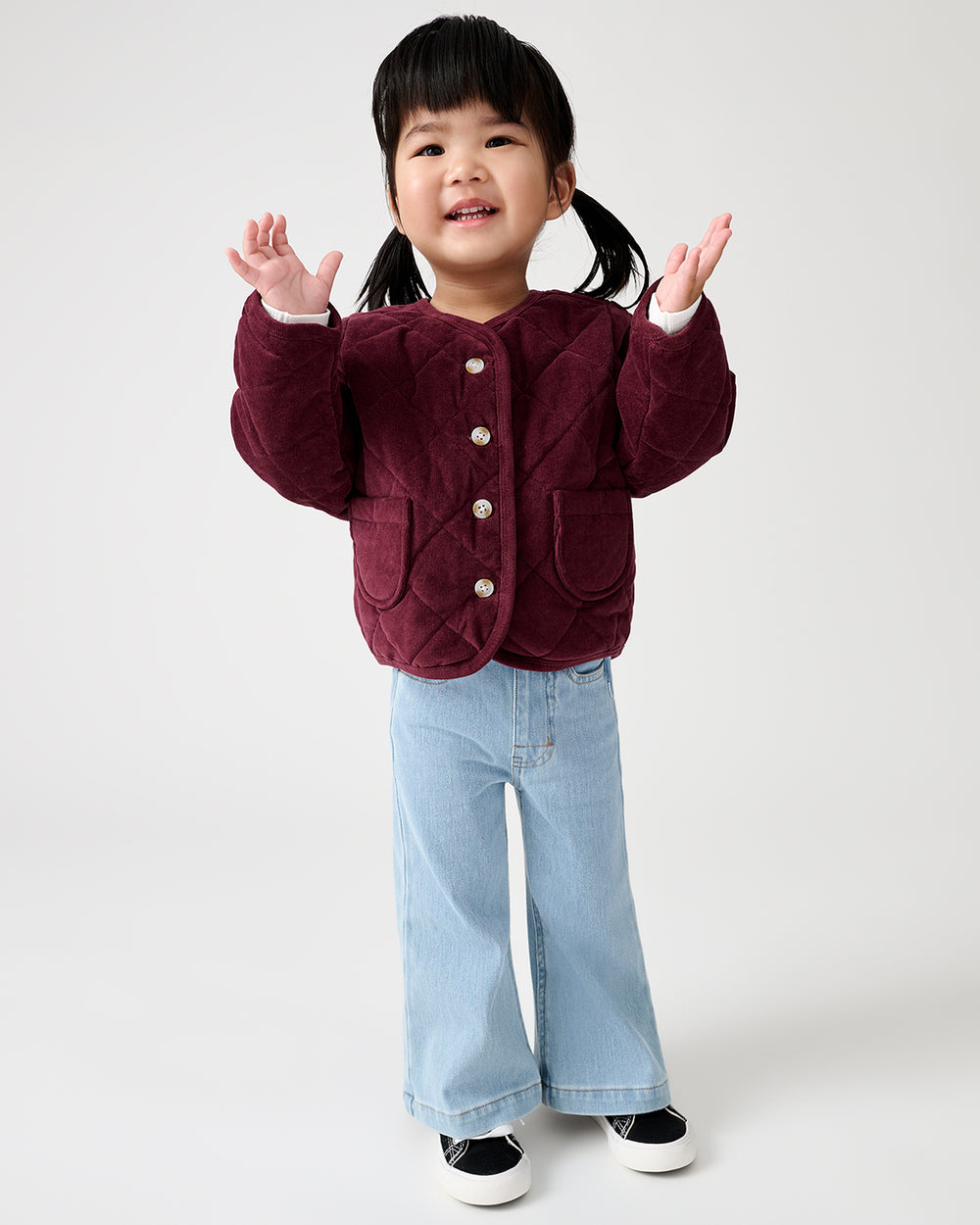 A young child with dark hair in pigtails stands against a light gray studio background, wearing a quilted burgundy jacket with buttons, light blue wide-leg jeans, and black-and-white sneakers.