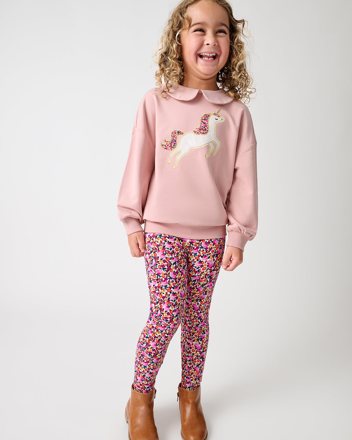 A young girl with curly blonde hair smiling and posing in a studio. She is wearing a light pink crewneck sweatshirt with a white and pink unicorn graphic on the front, paired with pink, purple, and yellow floral print leggings, and brown ankle boots.