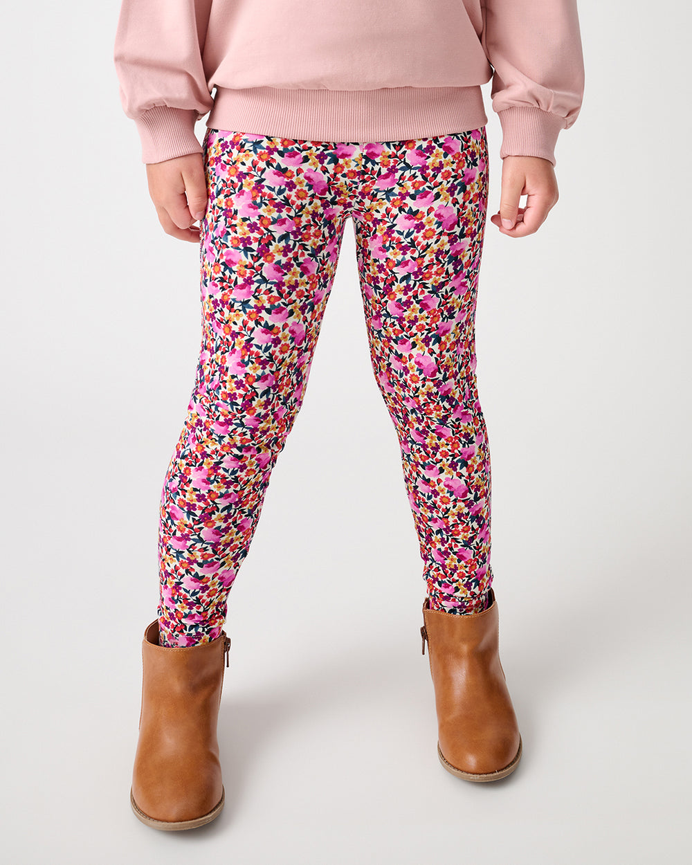 A child wearing pink floral leggings with a multicolor abstract pattern, styled with a light pink sweatshirt and brown ankle boots, shown from the waist down on a light gray studio background.