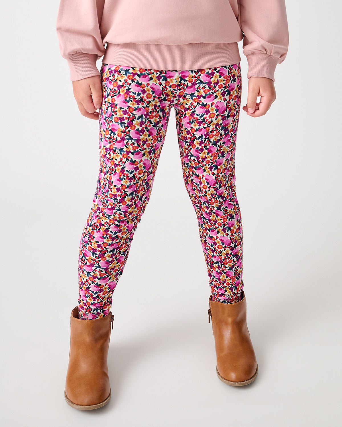 A child wearing pink floral leggings with a multicolor abstract pattern, styled with a light pink sweatshirt and brown ankle boots, shown from the waist down on a light gray studio background.