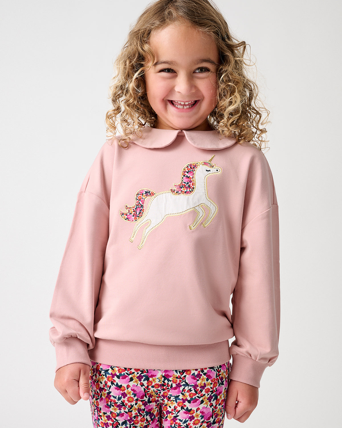 A young girl with curly blonde hair smiling, wearing a dusty pink sweatshirt with a white and pink unicorn applique on the front, paired with colorful floral pants, against a light gray studio background.