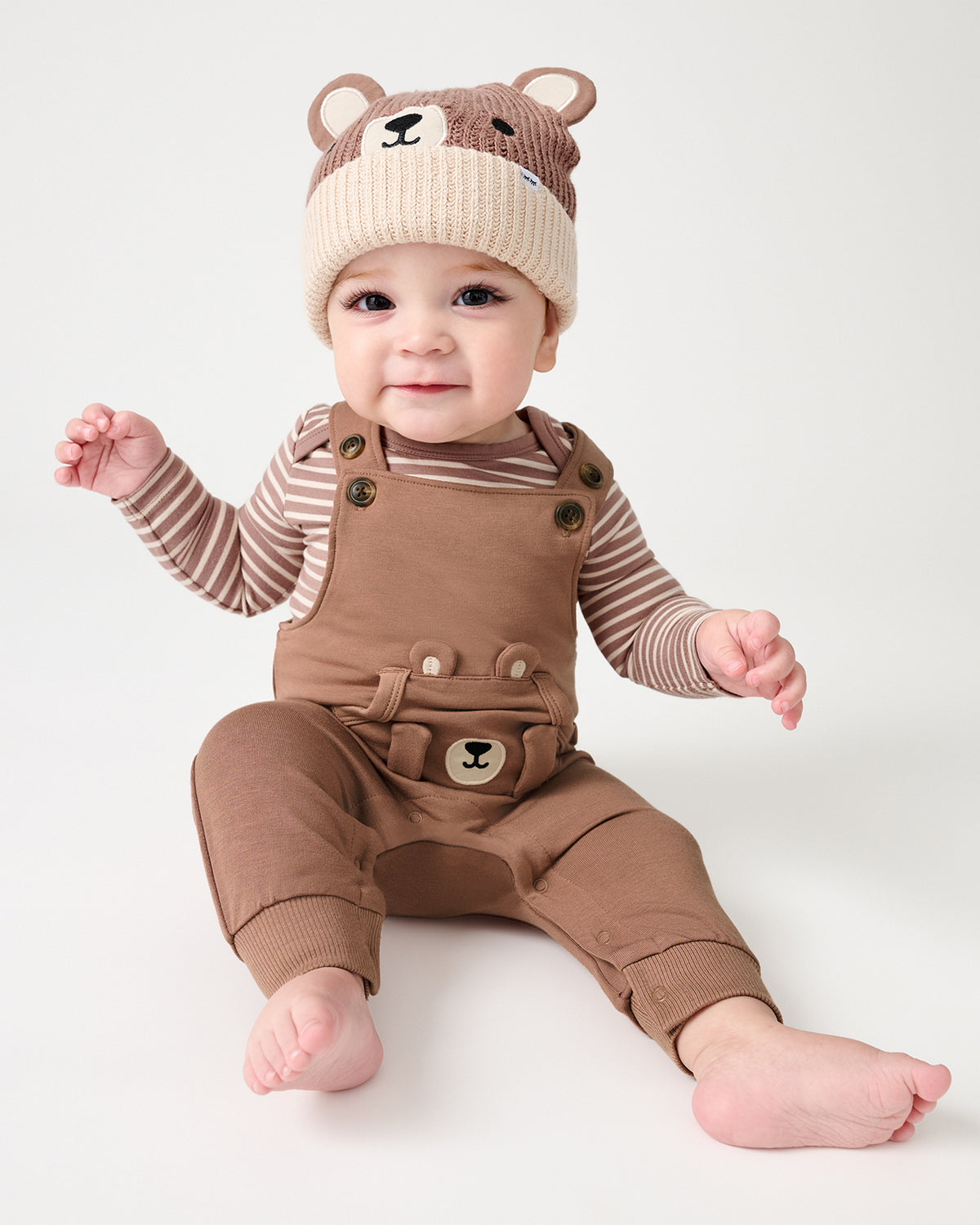 A baby sitting on a light gray studio background wearing a brown overall outfit with a matching beanie hat featuring a bear face.
