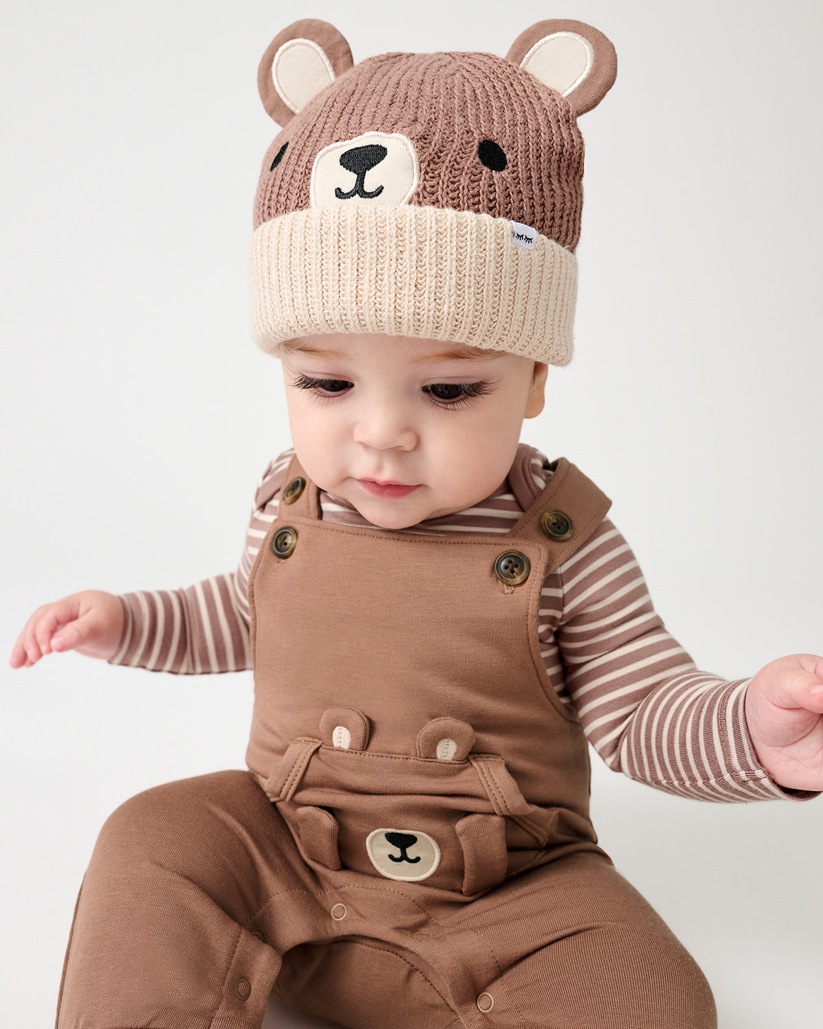 A baby wearing a brown knit bear-face beanie and brown overall with a striped long-sleeve shirt, photographed against a plain light background.
