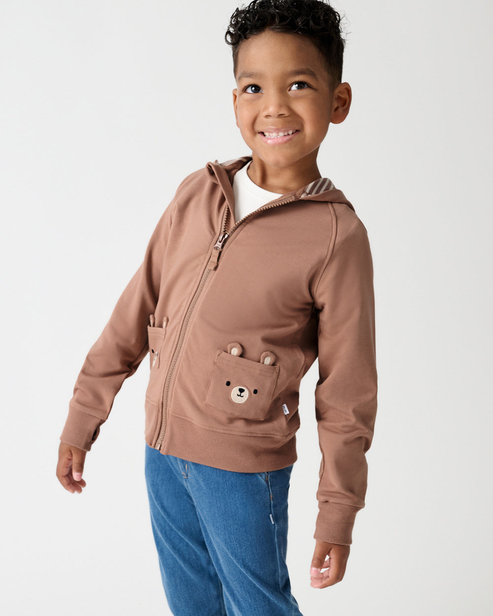 A young boy wearing a brown zip-up hoodie with a bear face pocket detail and blue jeans, standing against a light gray studio background.
