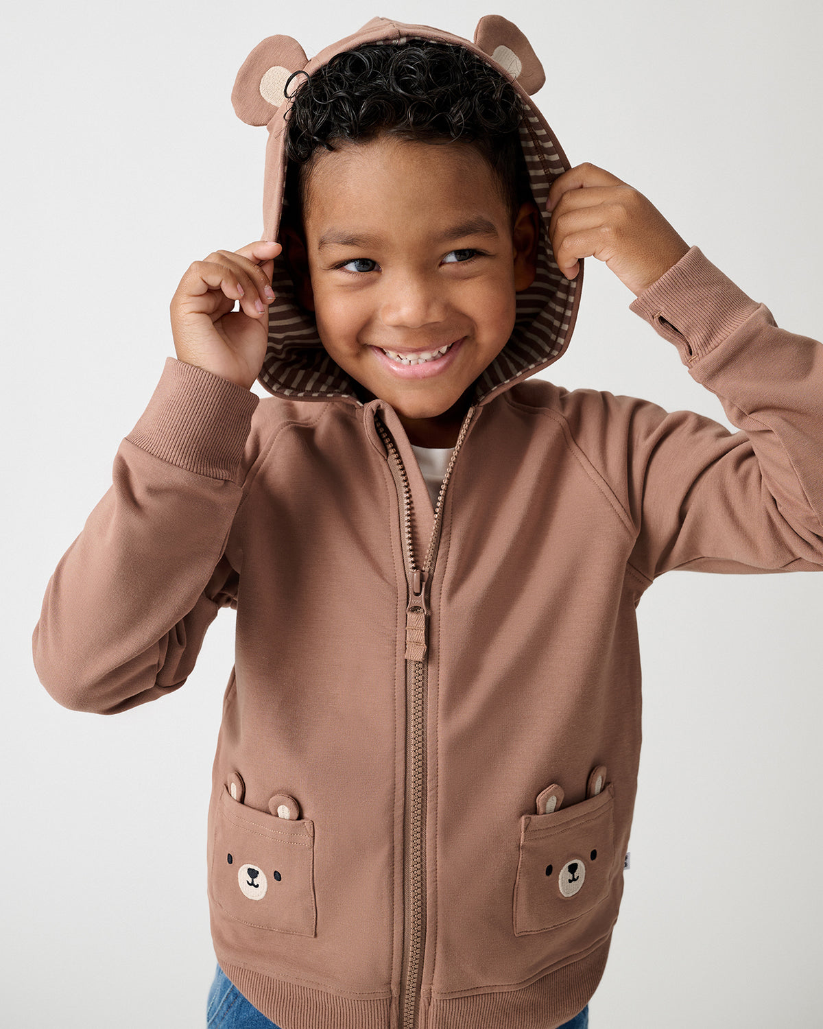 A smiling young boy wearing a brown zip-up hoodie with bear ears on the hood and bear face pockets, posing against a light gray background.