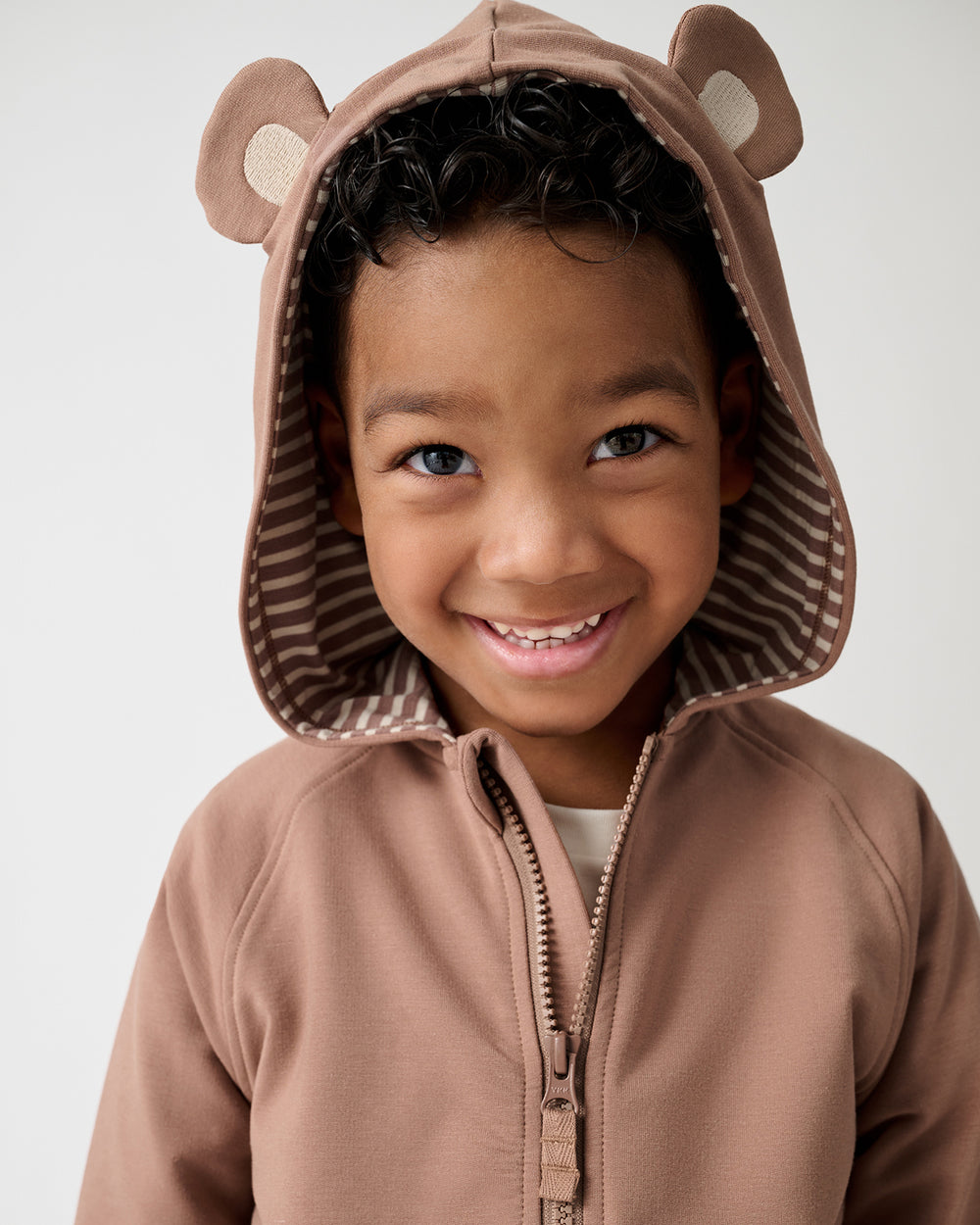 A smiling young child wearing a brown hooded zip-up outfit with bear ears on the hood, photographed against a clean light gray studio background.