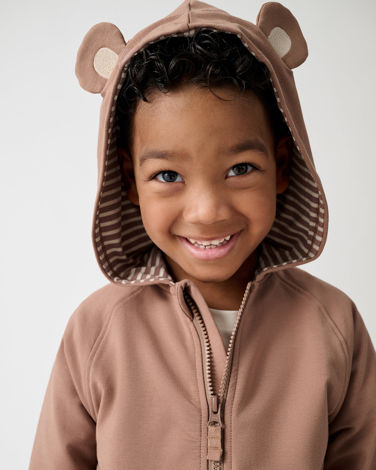 A smiling young child wearing a brown hooded zip-up outfit with bear ears on the hood, photographed against a clean light gray studio background.