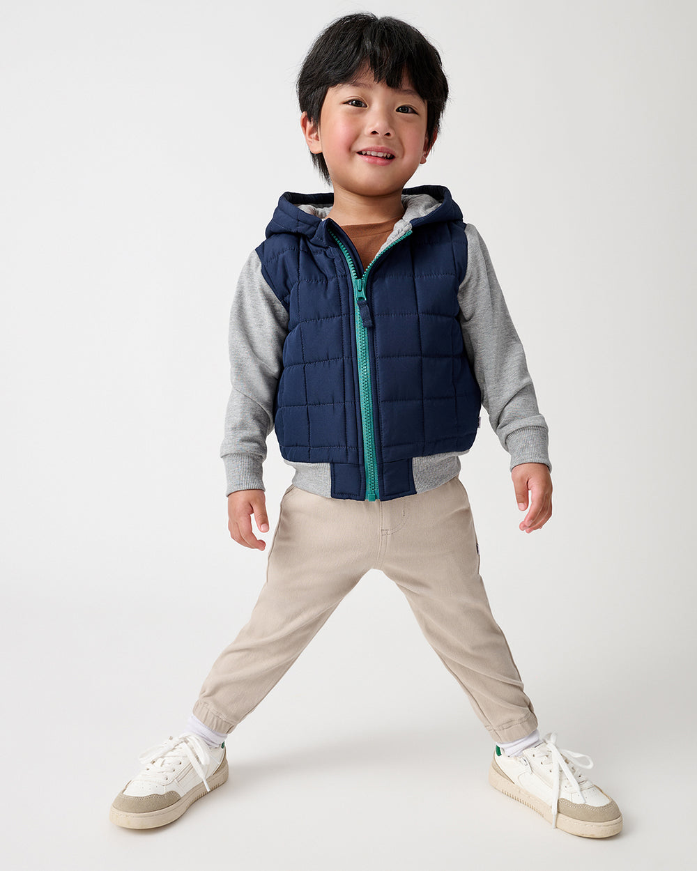 A young boy modeling a navy blue hooded jacket with gray long-sleeves, paired with beige pants and white sneakers, against a light gray studio background.