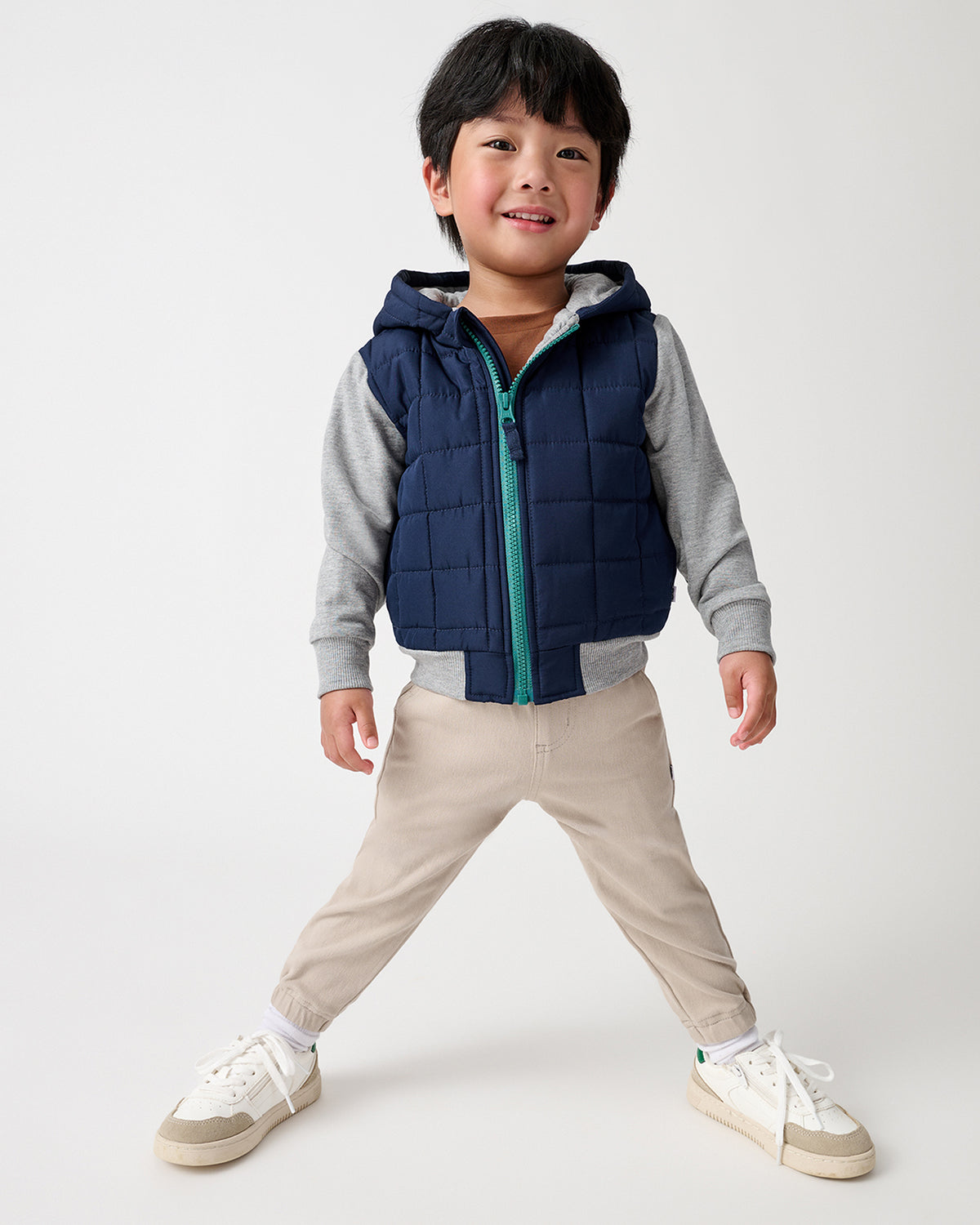 A young boy modeling a navy blue hooded jacket with gray long-sleeves, paired with beige pants and white sneakers, against a light gray studio background.