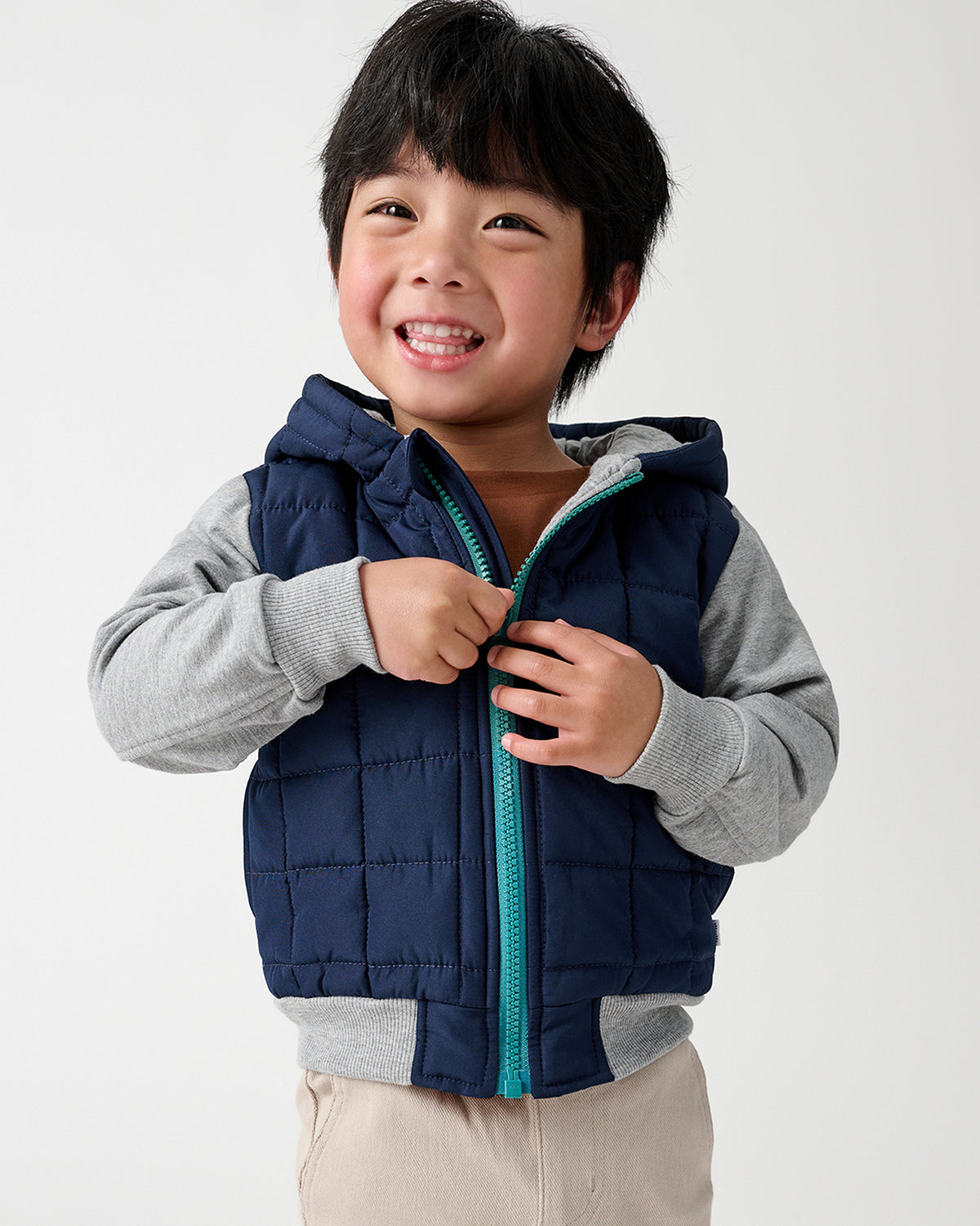 A smiling young boy wearing a navy blue hooded puffer jacket with gray sleeves and beige pants, holding the front zipper closed against a plain light gray studio background.
