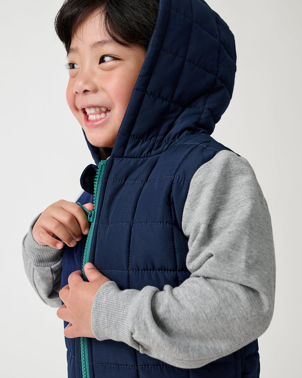 A smiling young boy wearing a navy blue quilted hooded jacket with heather gray long-sleeves, holding the zipper with a teal pull tab, against a plain light gray background.