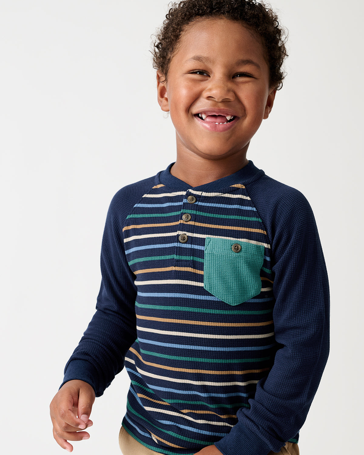 A smiling young boy wearing a long-sleeve raglan henley shirt with navy blue sleeves and a striped chest panel in navy, teal, beige, and white, featuring a teal chest pocket.