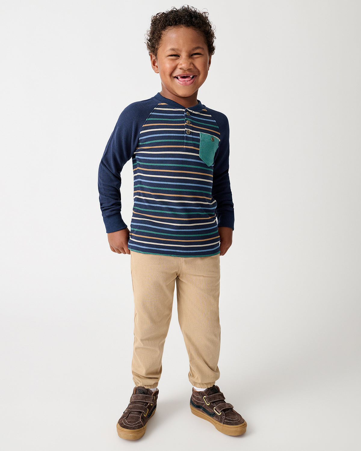 A young boy smiling and posing in a navy blue long-sleeve raglan henley shirt with thin horizontal stripes, beige pants, and brown shoes against a light gray studio background.
